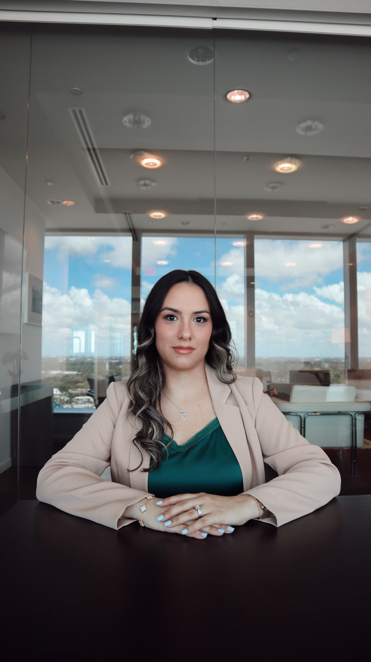 A woman with long wavy dark hair and light skin, wearing a beige blazer over a dark green top, sitting at a table in a modern office with large windows showing a cloudy sky, with her hands folded on the table.