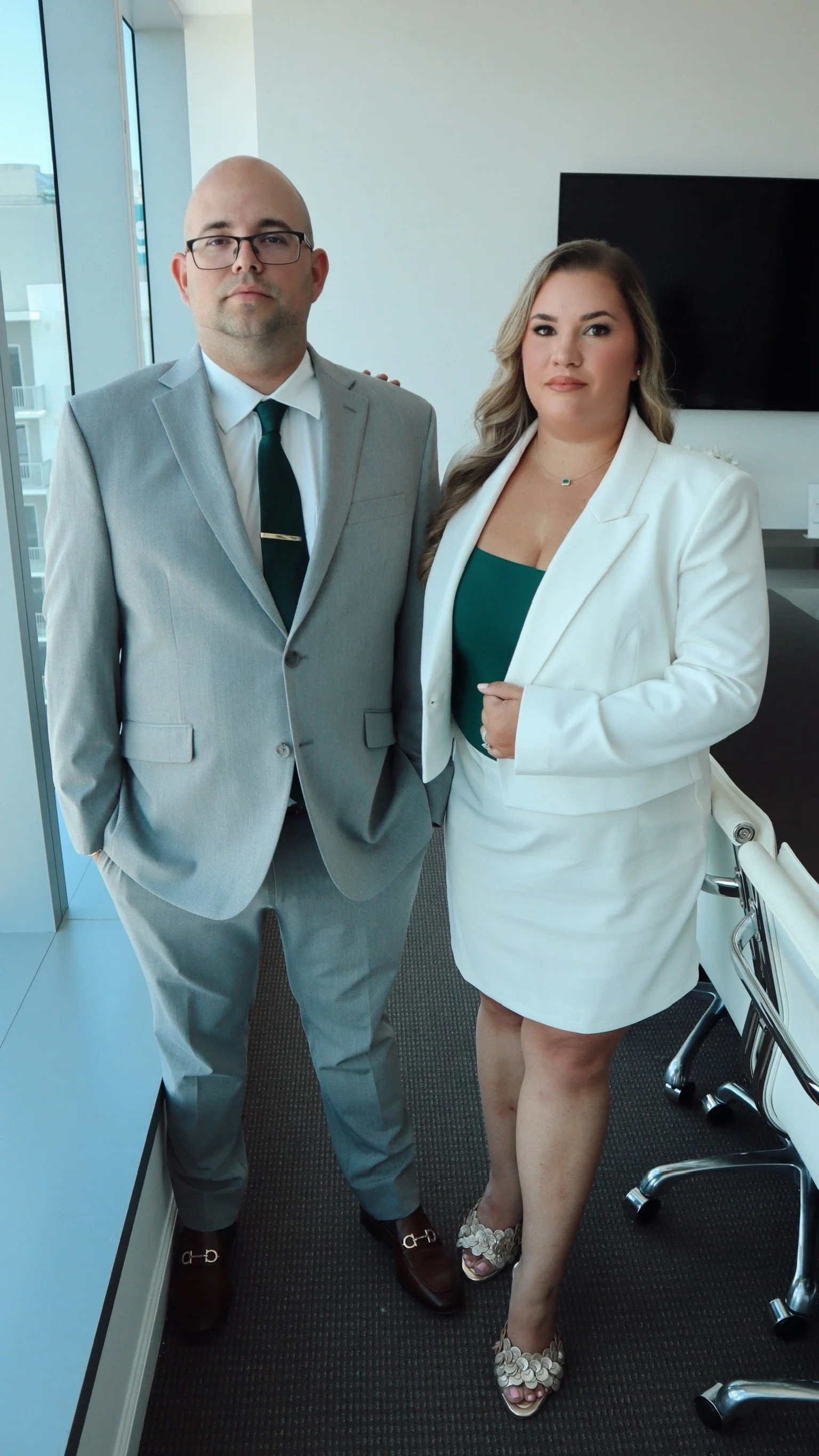 A male attorney and a female attorney standing in an office, dressed in business attire, with large windows and a television in the background.