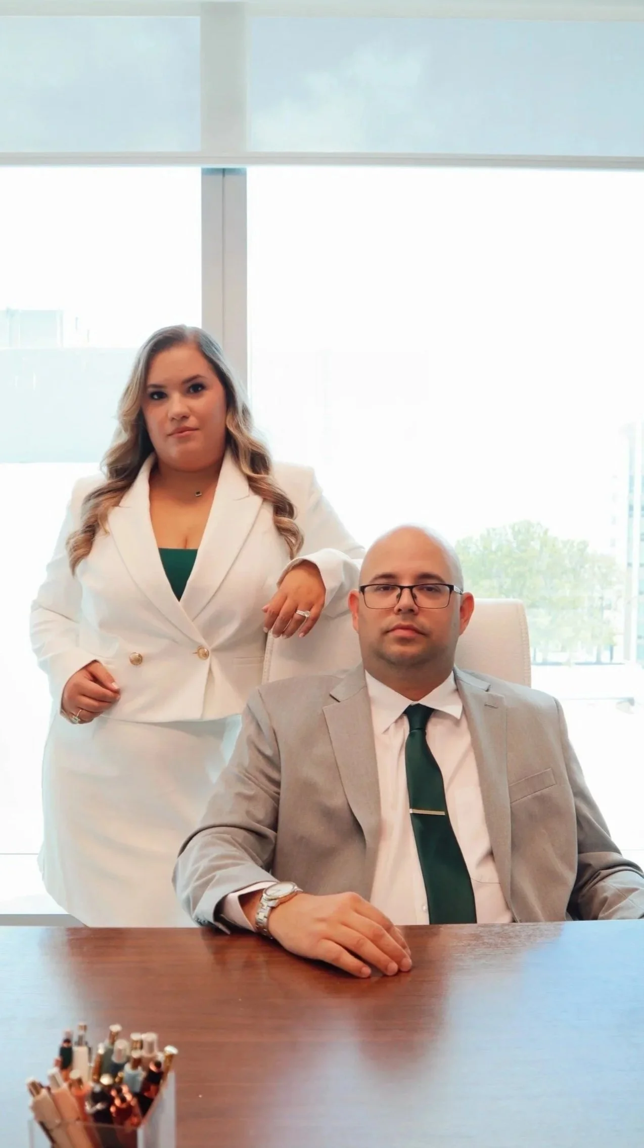 A female attorney standing behind a seated male attorney in a modern office, with large windows and a city view in the background.