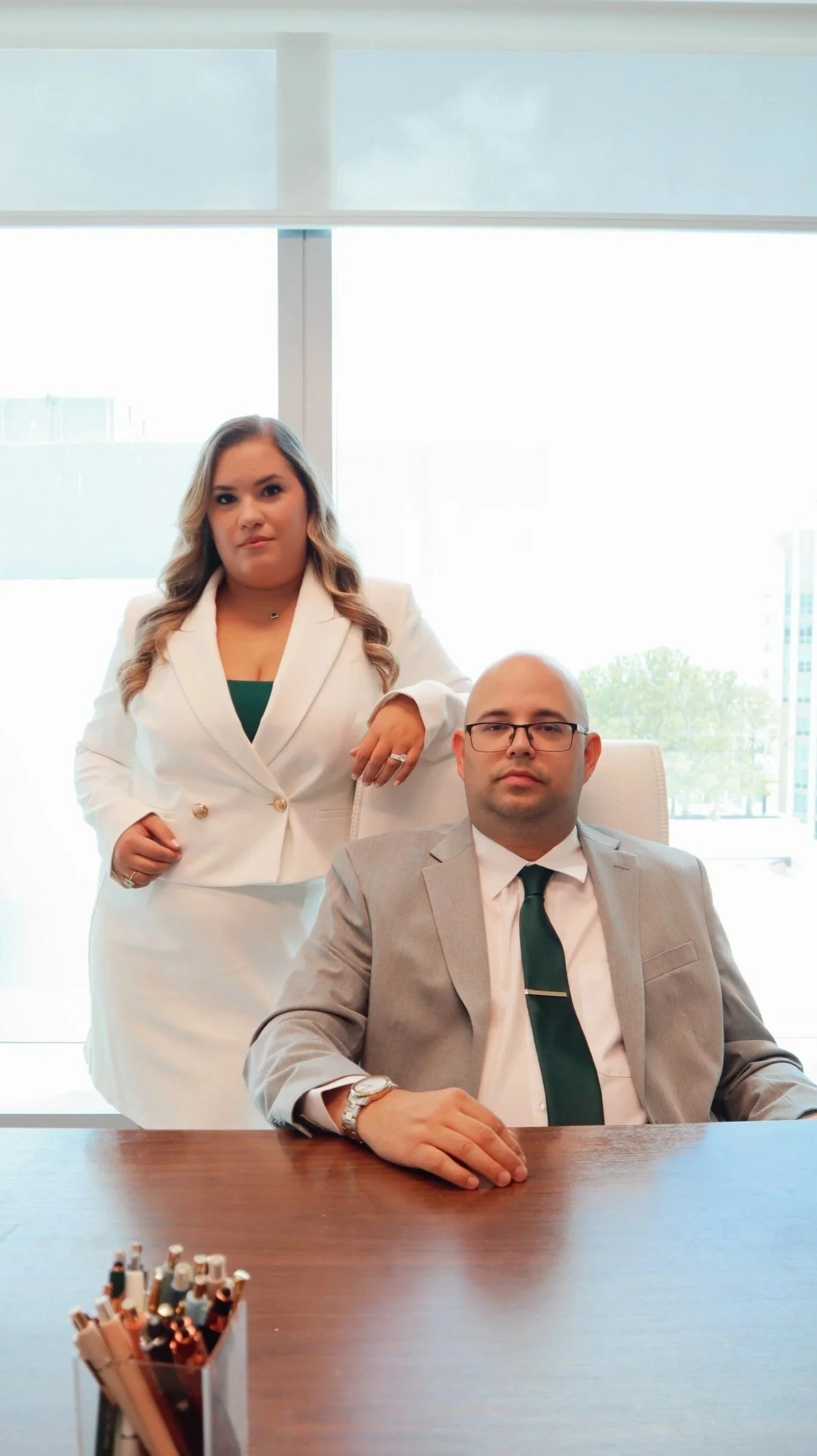 A female attorney in a white blazer standing next to a male attorney in a gray suit with glasses, sitting at a desk in an office with large windows and city buildings outside.
