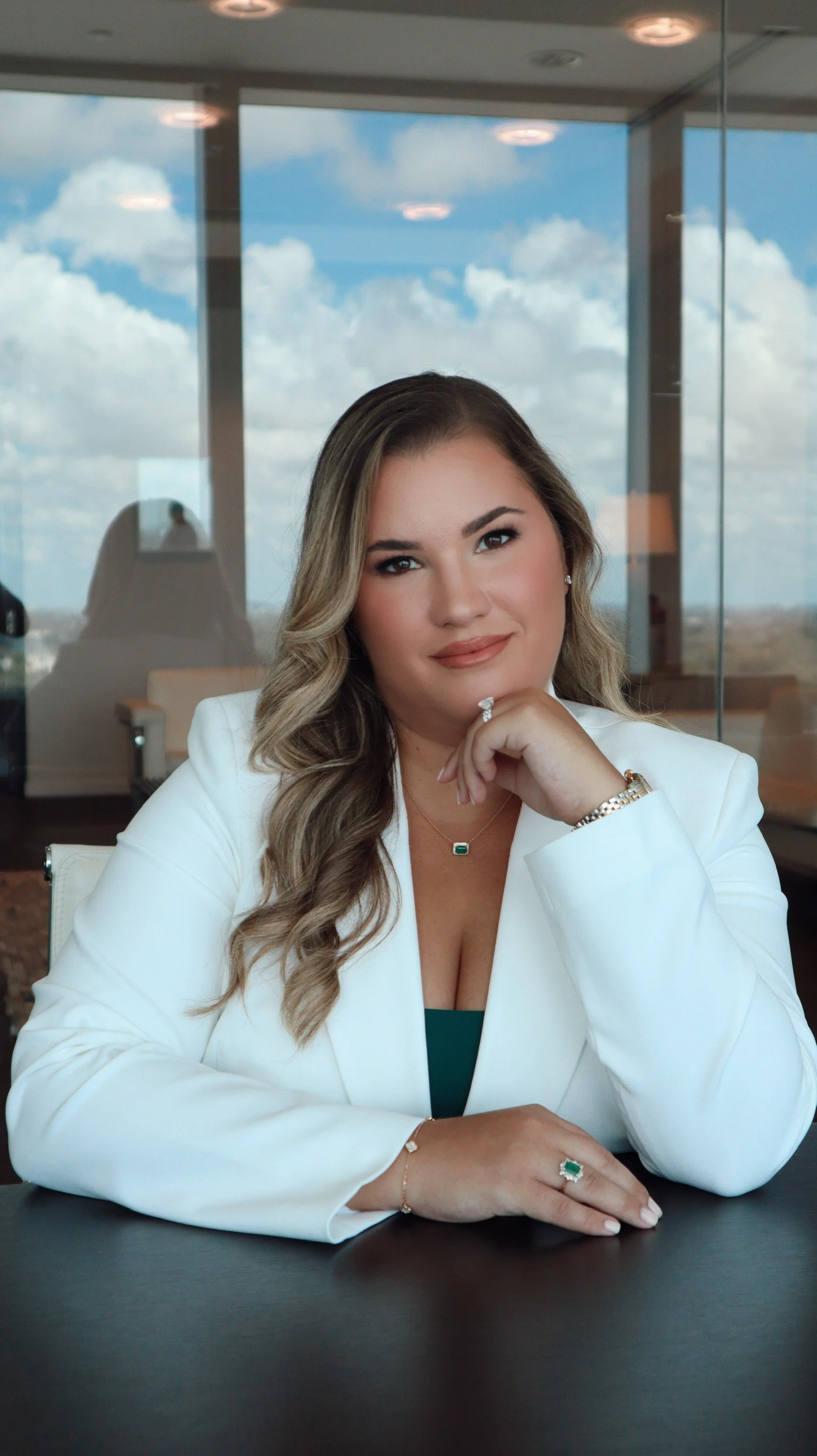 A female attorney sitting at a table in a modern office with a glass wall, large windows, and a city view. She has long wavy hair, is wearing a white blazer, and has jewelry, including a ring, bracelet, necklace, and earrings.