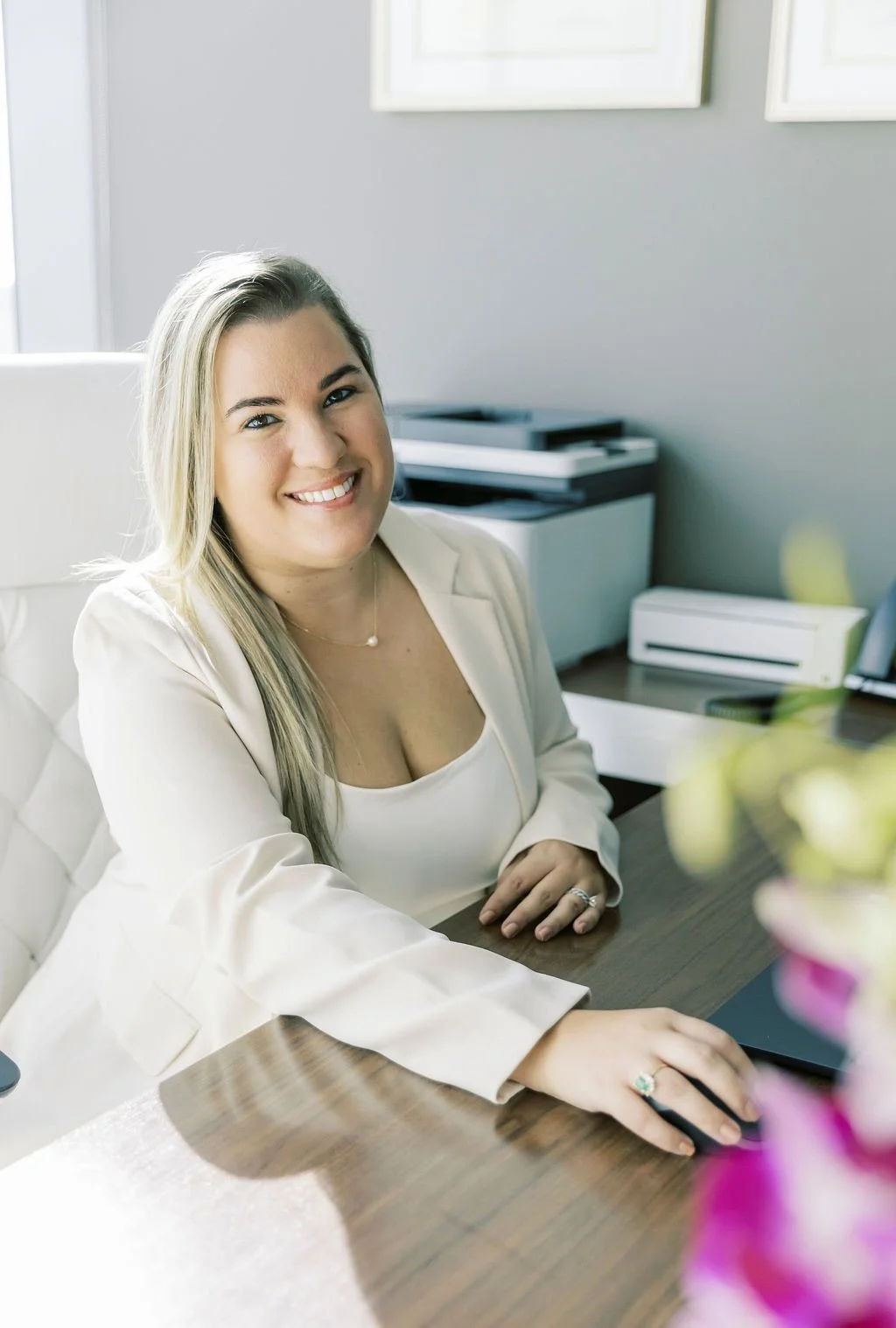 A smiling woman in a white blazer sitting at a desk with a computer mouse, in an office setting with framed pictures on the wall and a printer in the background.