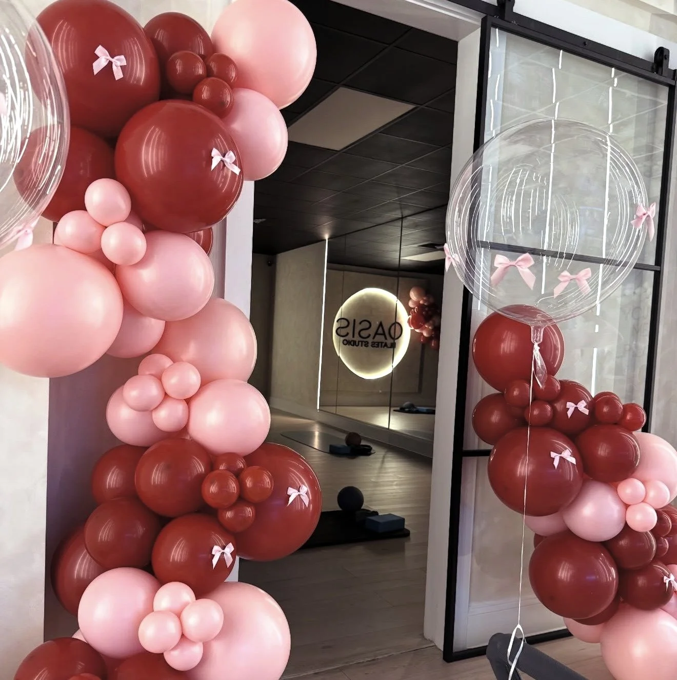 Decorative arch made of pink and red balloons with small pink bows, at the entrance of a room with a mirror reflecting the balloons. Inside the room, a sign reads 'OASIS' and there is a fitness mat with small weights.
