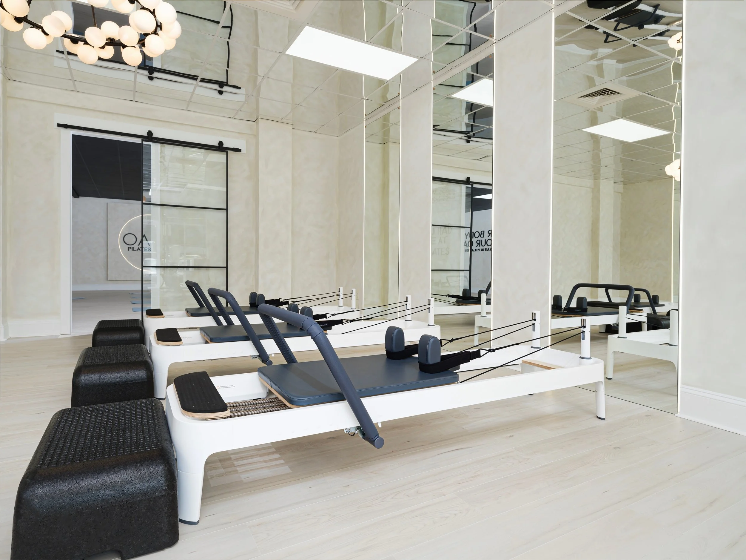 Fitness studio with mirrors on the wall, light-colored wooden floors, and workout equipment including mats, benches, and Pilates reformer machines. There is a chandelier with multiple round white bulbs hanging from the ceiling.