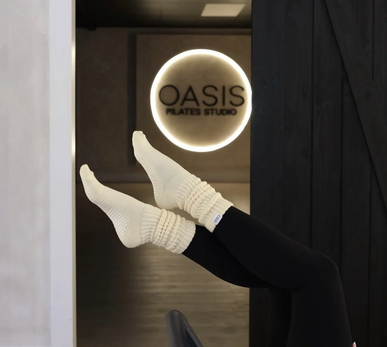Person lying on their back with legs raised, wearing white socks and black leggings in front of a glowing sign that reads 'OASIS FLAVES STUDIO' in a modern interior.