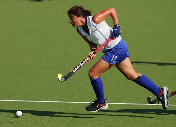 Female field hockey player in blue uniform and white jersey running on green turf with hockey stick chasing a ball.