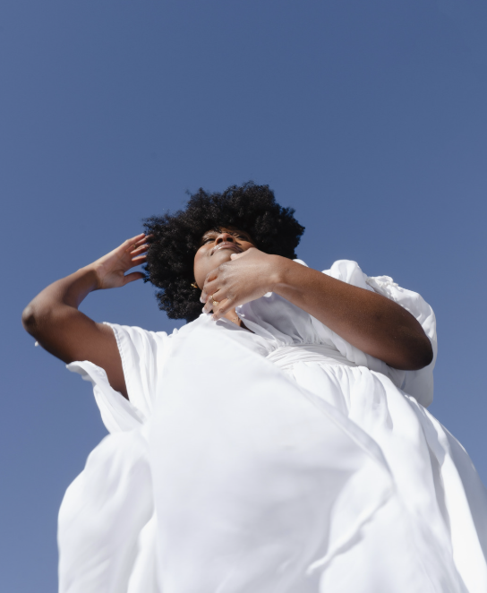 A woman with curly hair wearing a white dress standing against a clear blue sky.