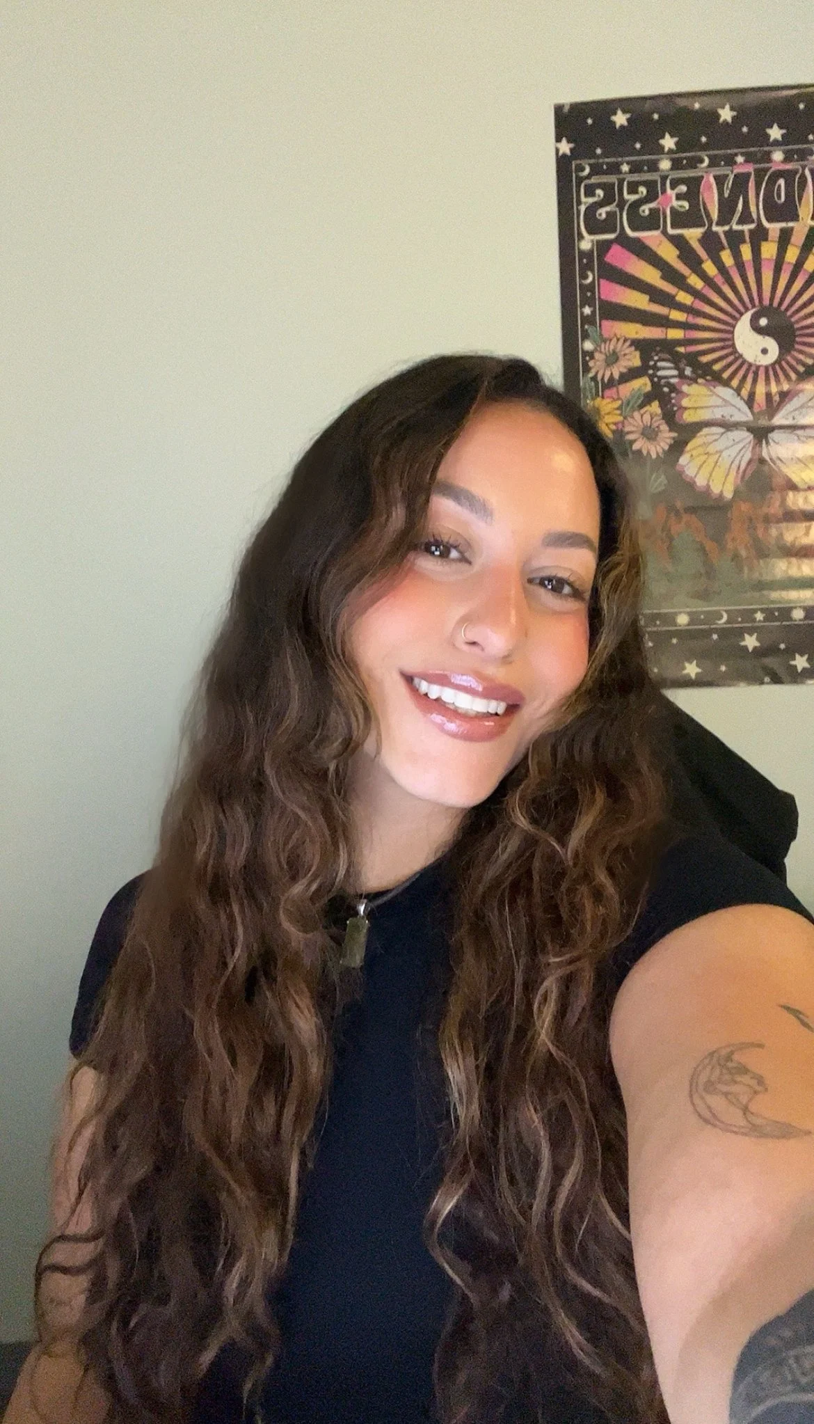 A woman with long, wavy brown hair and a nose ring, smiling at the camera, wearing a black shirt, with a colorful art poster featuring spiritual symbols and motifs in the background.