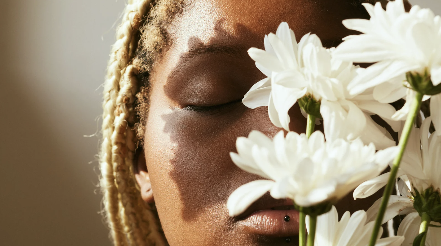 Close-up of a woman with blonde braids, her eyes closed, holding white flowers close to her face, with sunlight creating shadows on her skin.