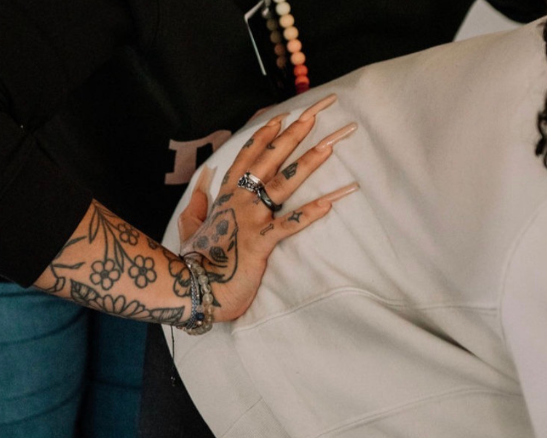 A person with long, manicured nails resting their hand on another person's white clothing. The person has multiple tattoos including flowers on their arm and a skull on their hand, and wears rings and bracelets.