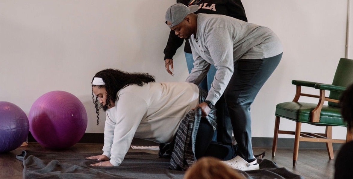 A woman practicing yoga or stretching on a mat with assistance from a man, with exercise balls and a chair nearby.