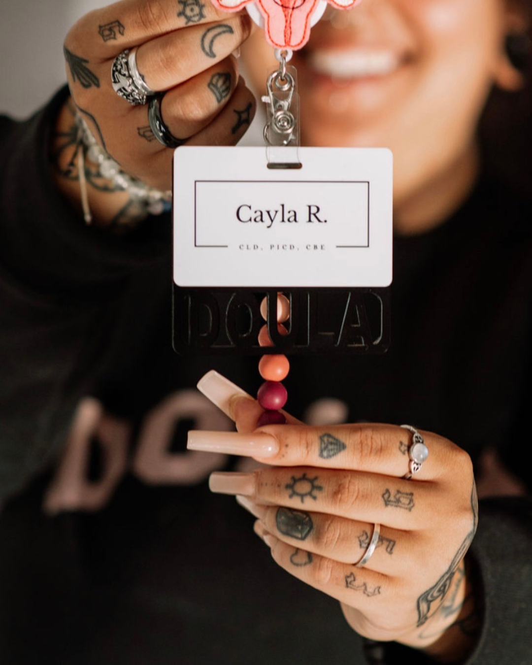 Person with multiple tattoos holding a conference or event badge with the name "Cayla R." and a lanyard with the word "DOLLA" visible. The person has long, manicured nails, rings, and various tattoos on their hands and fingers.