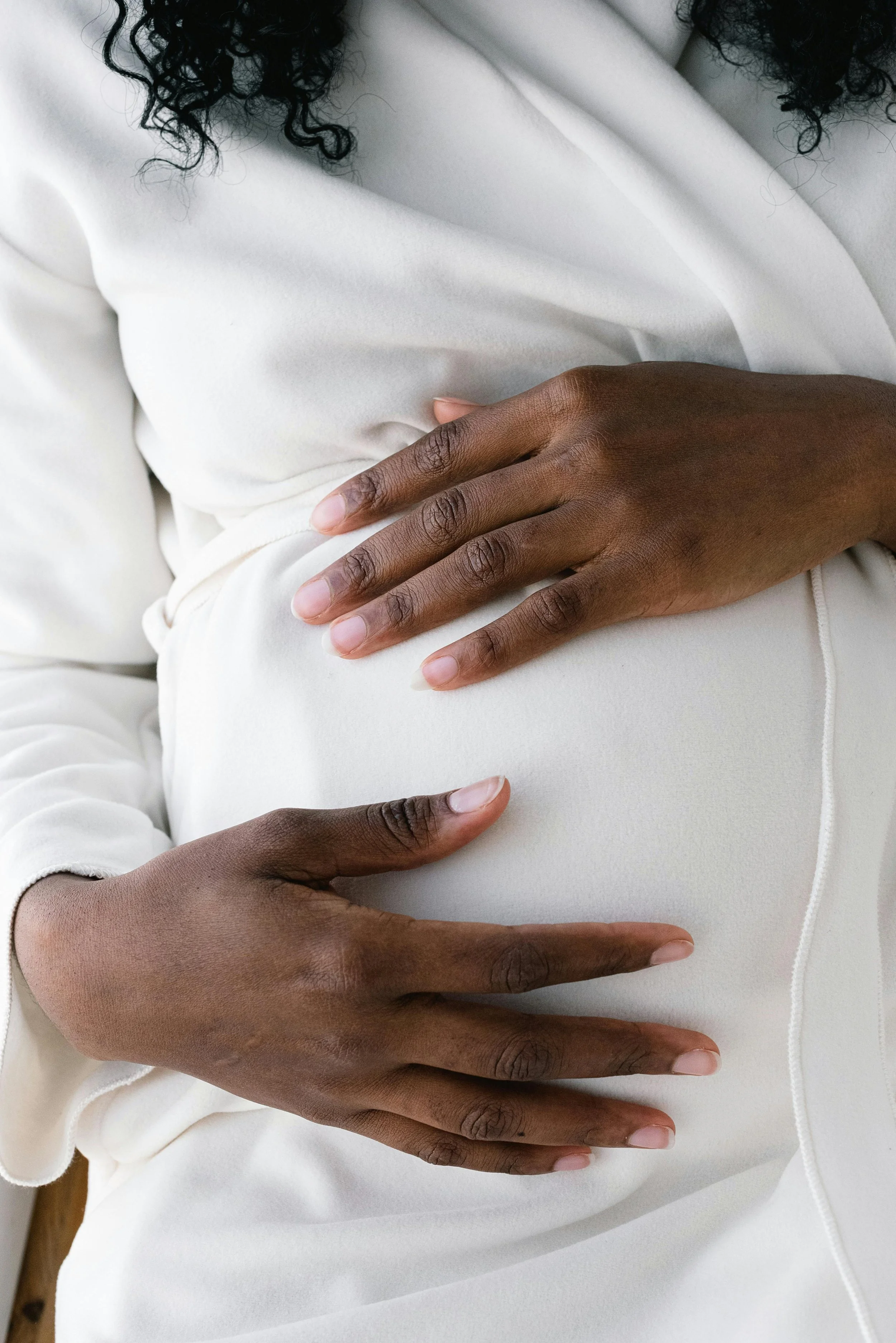 Close-up of a woman in white clothing gently resting one hand on her pregnant belly, her skin tone is dark, and her hair is curly and black.