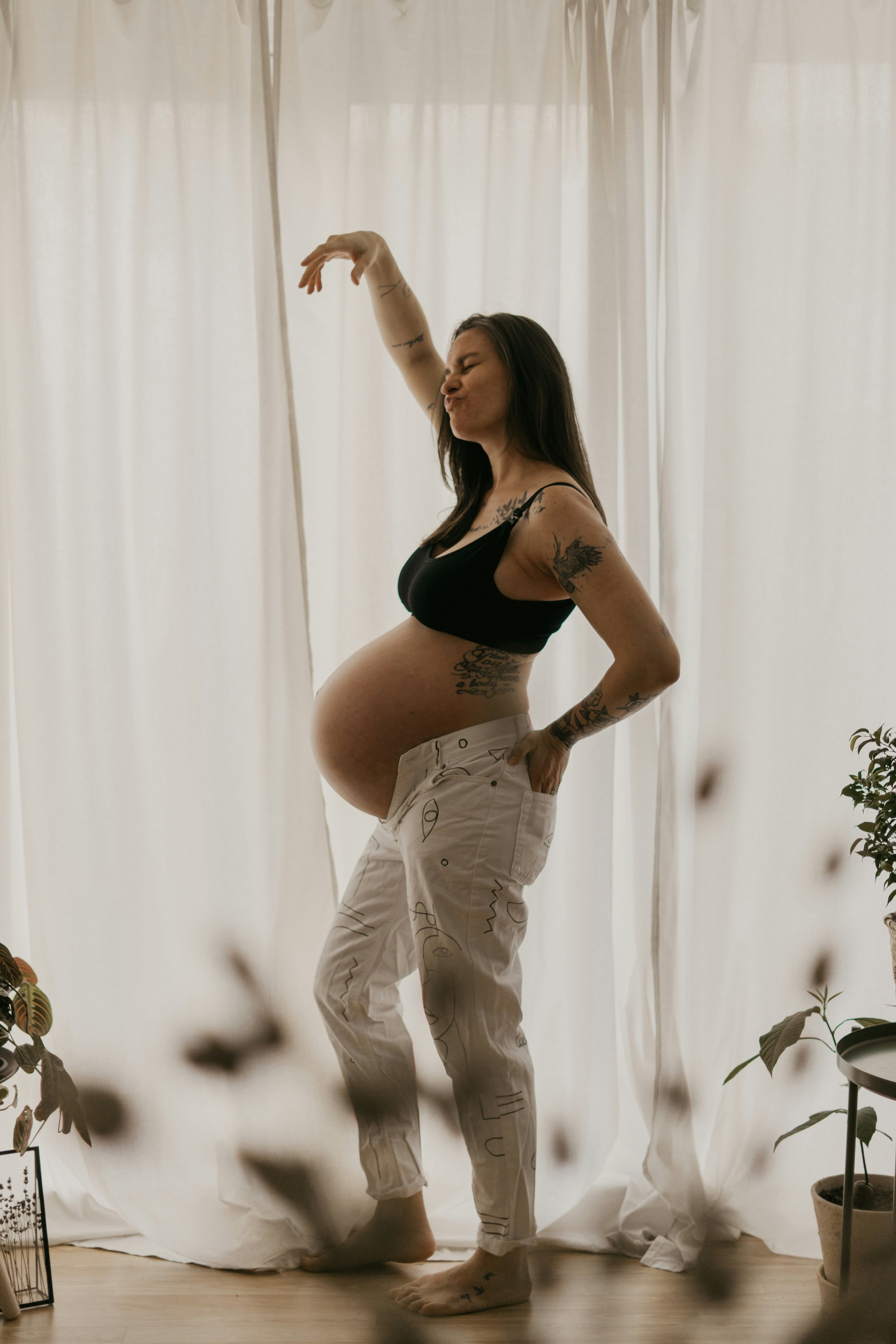 Pregnant woman with tattoos posing inside near white curtains, wearing black top and patterned beige pants.