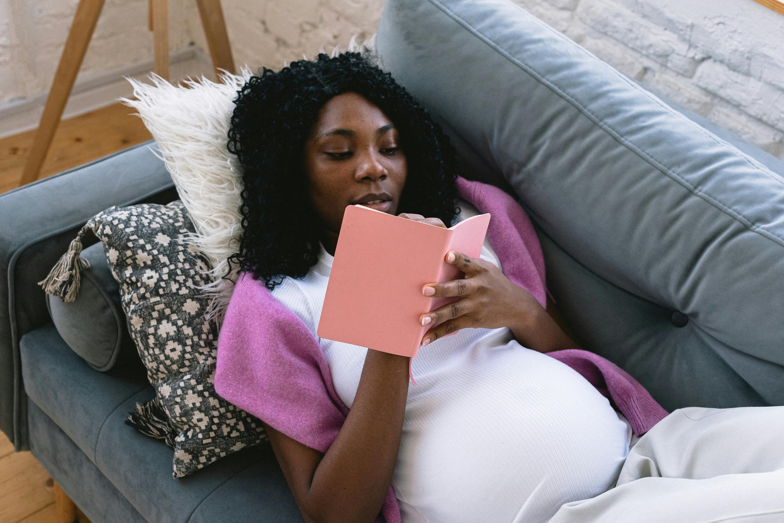 A pregnant woman with curly black hair lounging on a gray sofa, reading a pink book, with a decorative pillow behind her head, in a cozy home setting.