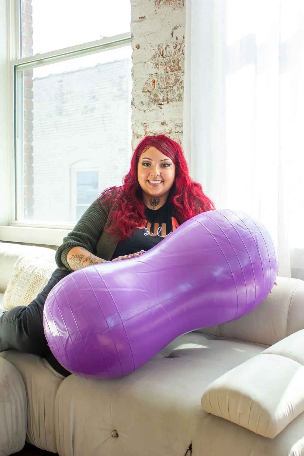 A woman with red hair and tattoos sitting on a white sofa, holding a large purple inflatable bean bag in a room with large windows and white walls.