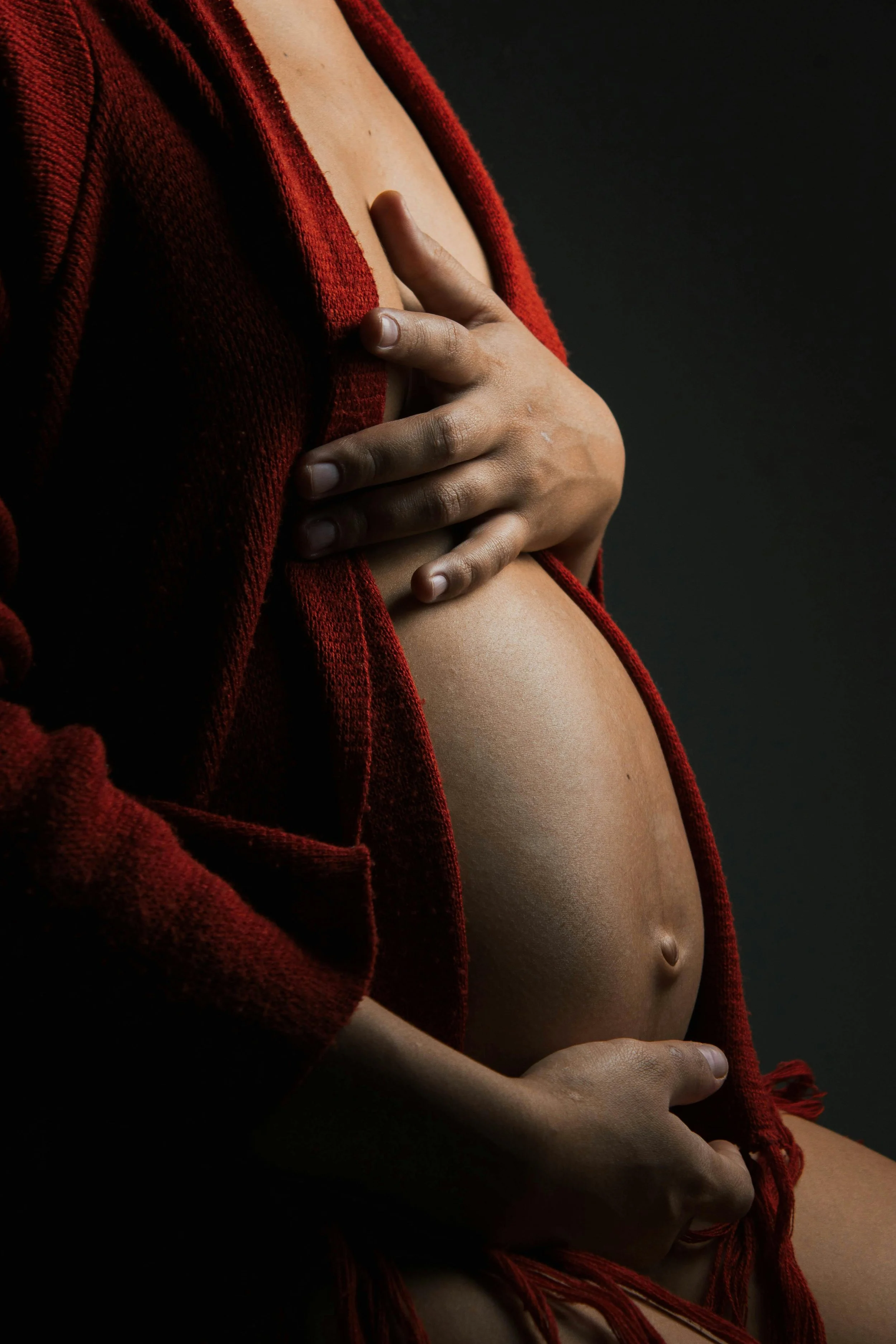 A pregnant woman with bare skin, wearing a red knitted sweater, her hand gently resting on her stomach.