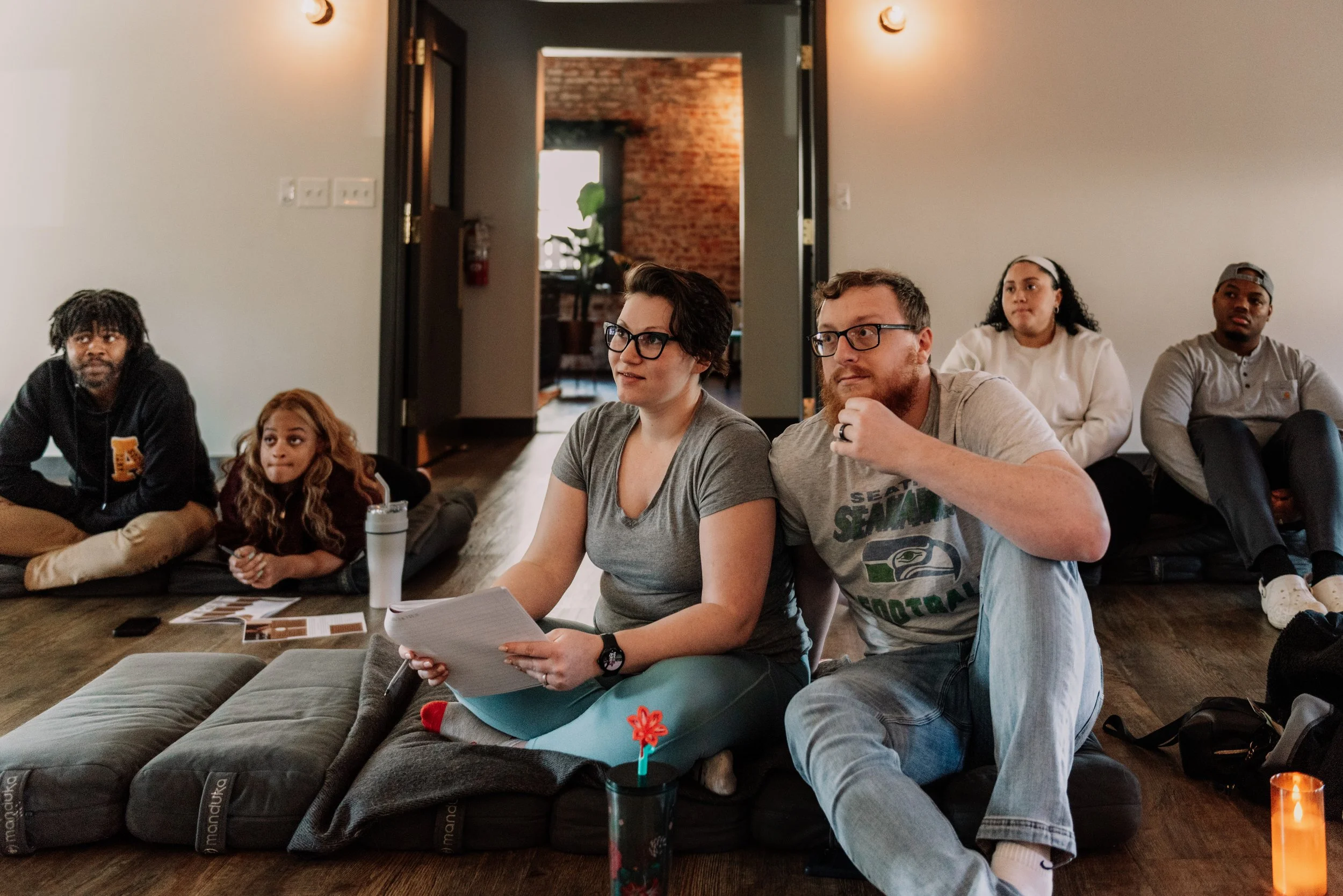 A group of people sitting on the floor and cushions in a room, paying attention to something outside the frame. Some have notebooks, and there are photo prints and a water bottle on the floor.