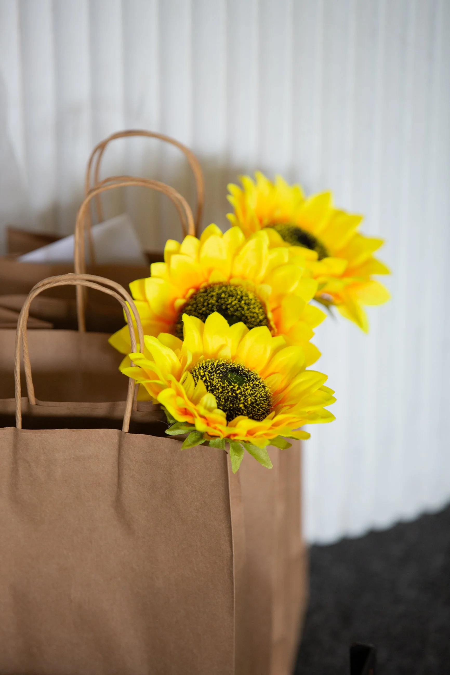 Yellow sunflower flowers in paper bags with handles on a wooden shelf against a white paneled wall. Guest VIP gift bags at a braless brunch event.