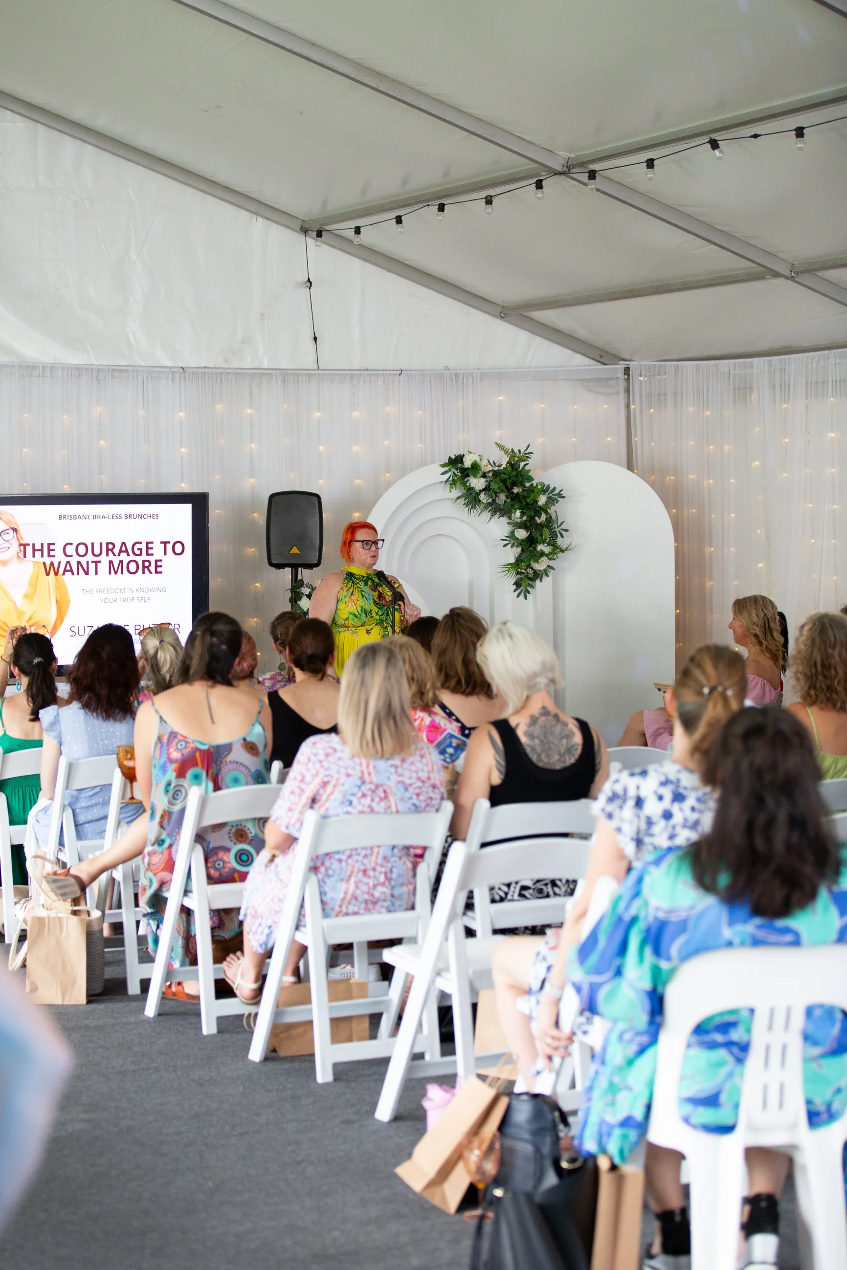 A woman in a yellow and green dress standing and speaking into a microphone at an event with a white backdrop decorated with a green floral arrangement. Audience members with various hairstyles and colorful clothing seated on white chairs.