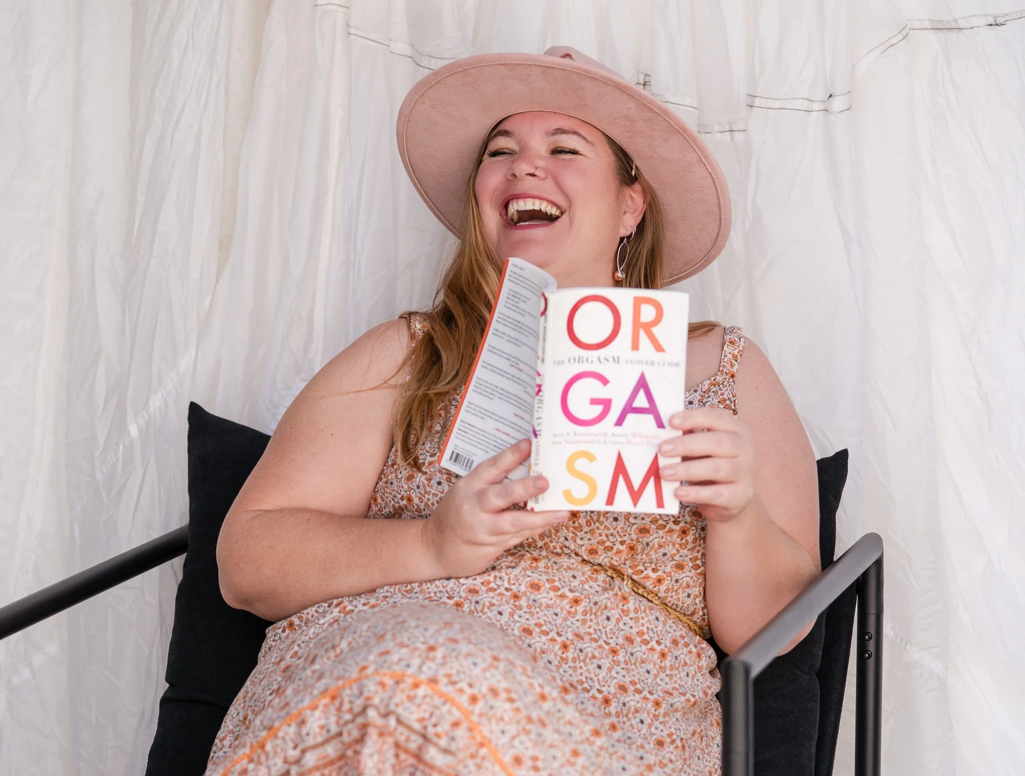 A woman with long red hair, wearing a large pink hat, earrings, and a floral dress, is sitting in a black chair, smiling while looking at a book titled 'OR,' with the subtitle partially visible, in front of a white curtain backdrop.