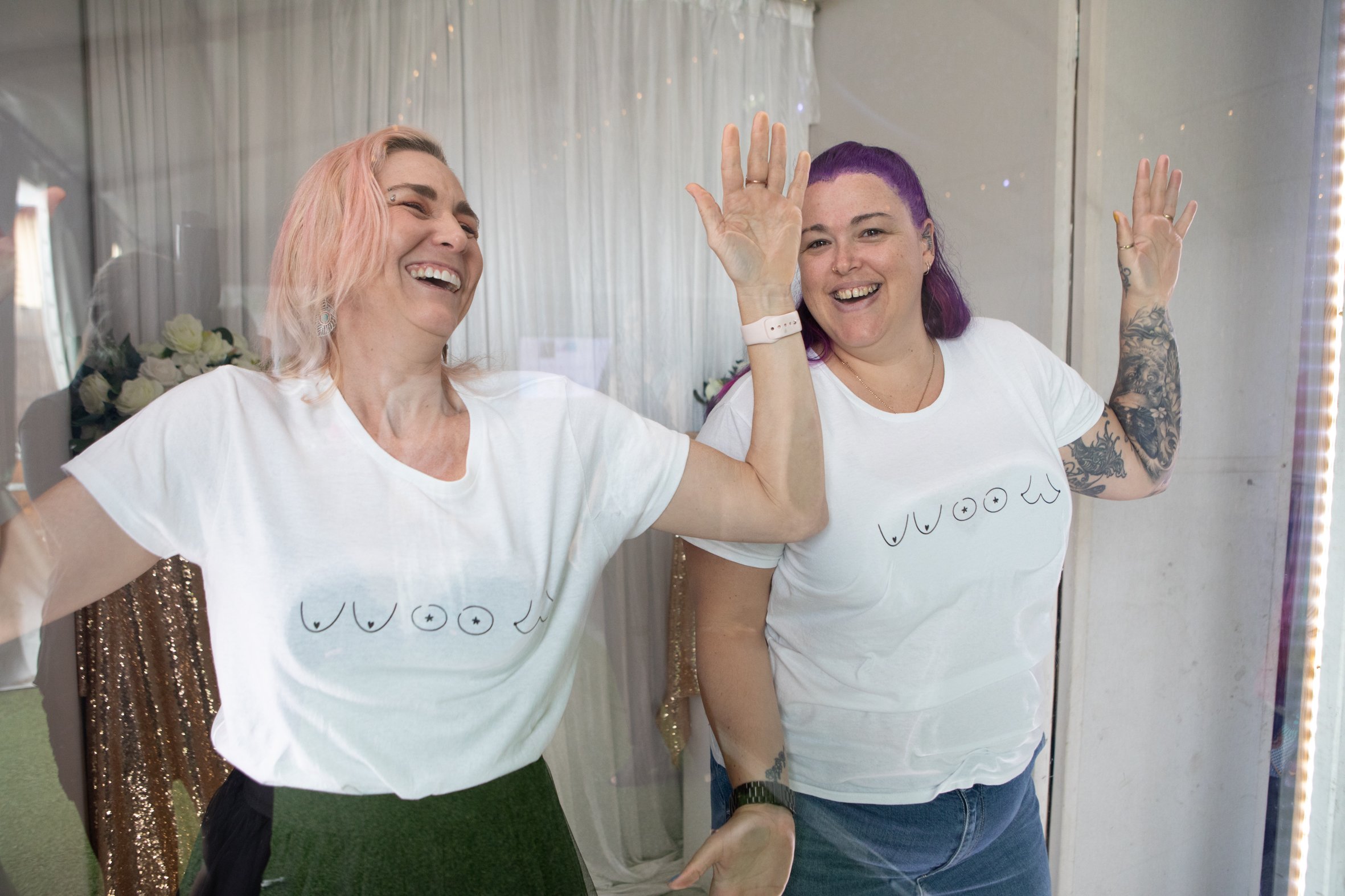 Two women wearing white t-shirts with printed designs are in front of a mirror, smiling and waving. One has short pink hair, the other has purple hair and tattoos on her arm. The environment appears festive with curtains and decorative lights in the background.