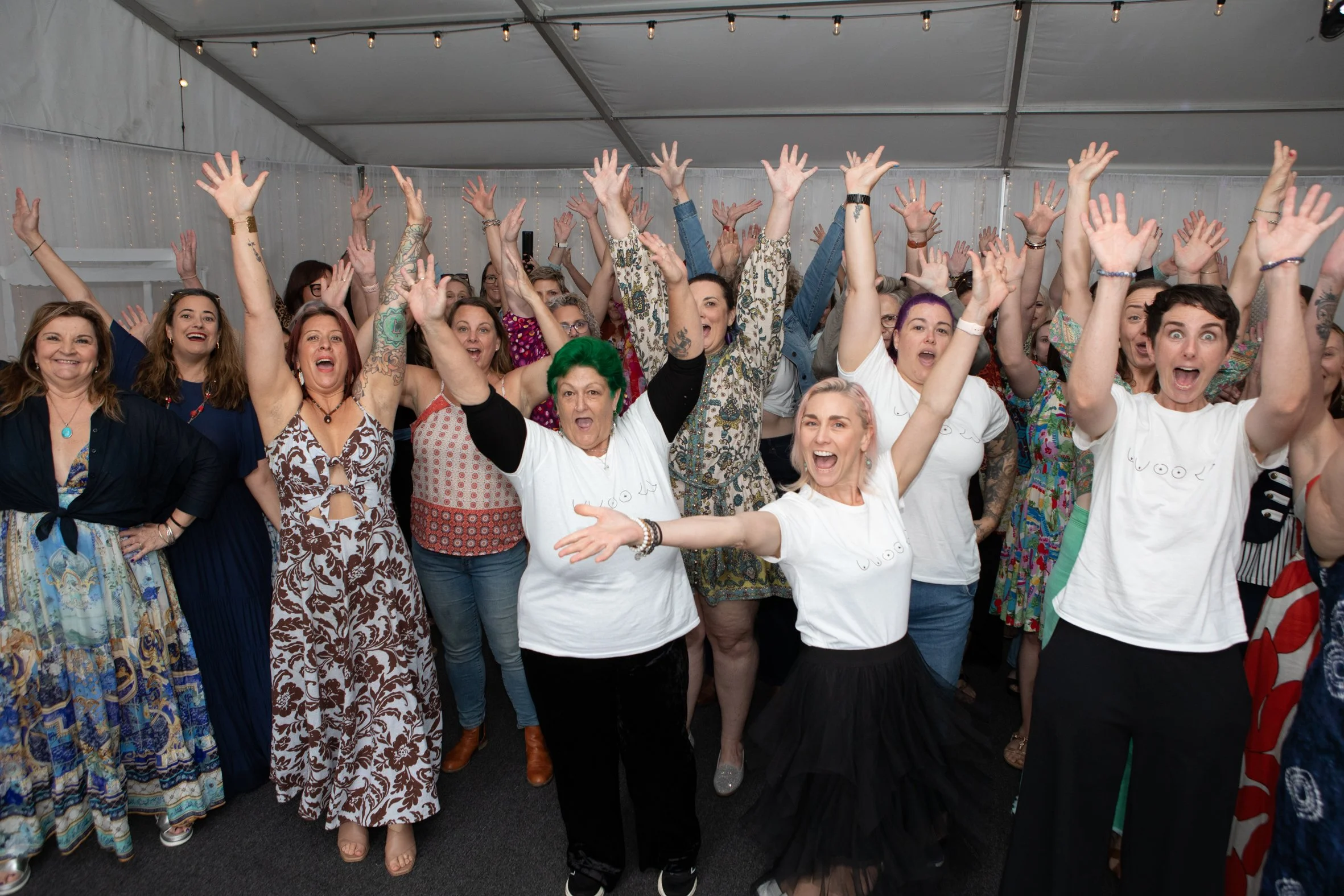 A large group of women indoors at brisbane Braless brunch event, celebrating with their hands raised and smiling.