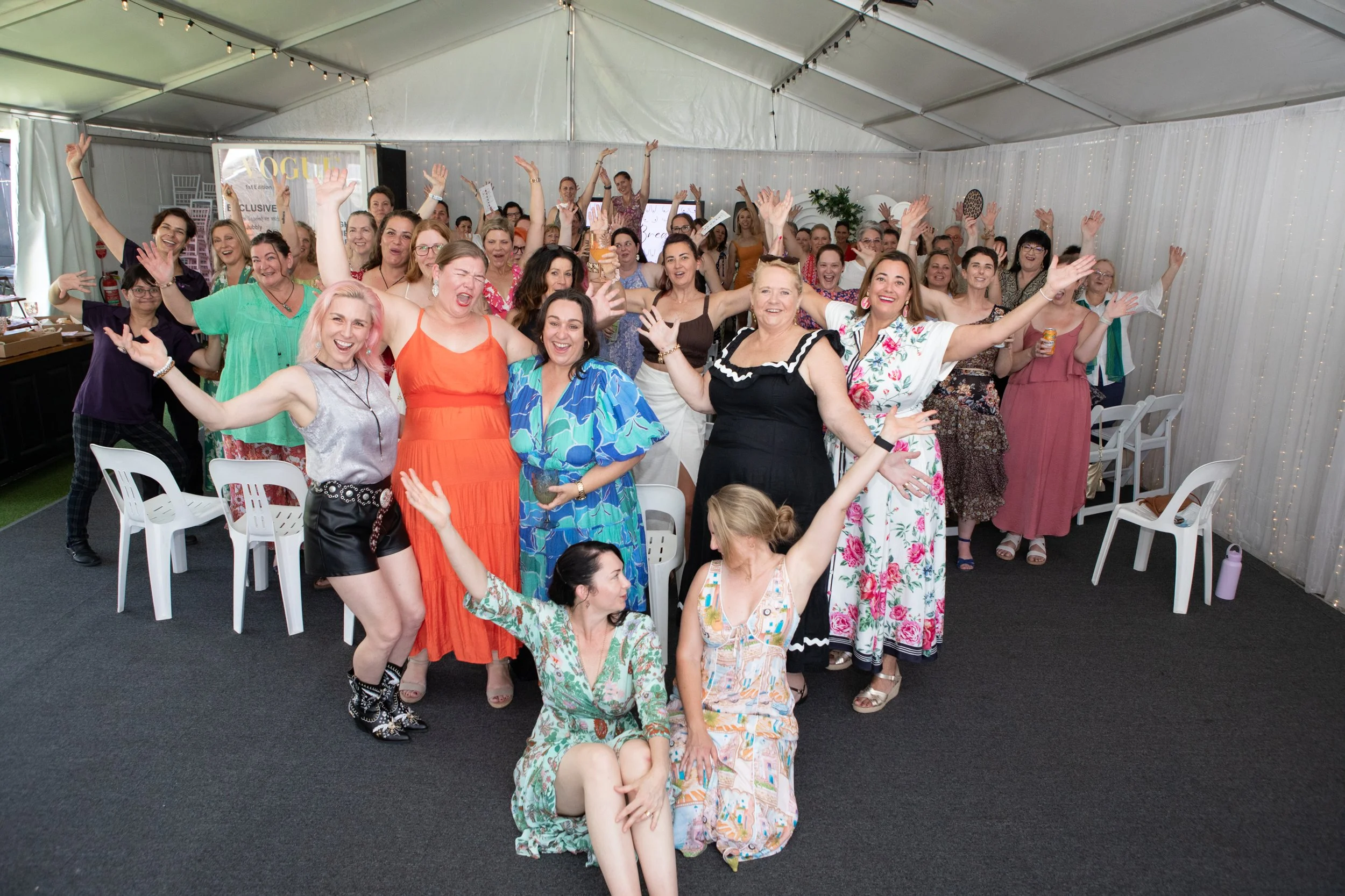 A large group of women at at Braless brunch event celebrating inside a decorated event tent, some standing and some sitting, with many raising their hands and smiling.