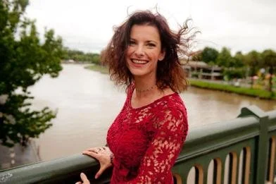 A woman with curly red hair wearing a red lace dress, smiling and standing by a green bridge rail over a river.