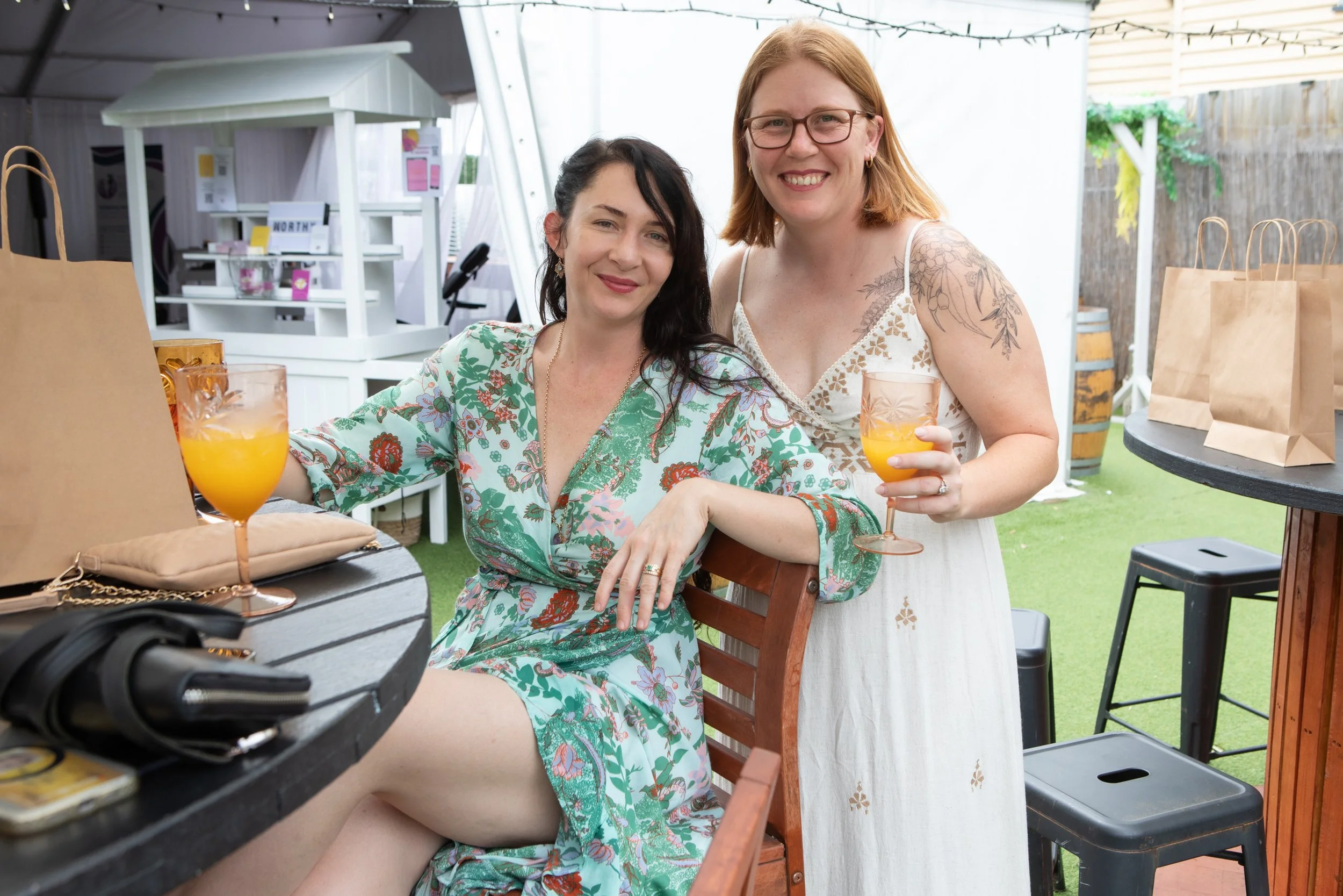 Two women smiling, sitting outdoors at a table with drinks, surrounded by a white tent and gift bags.