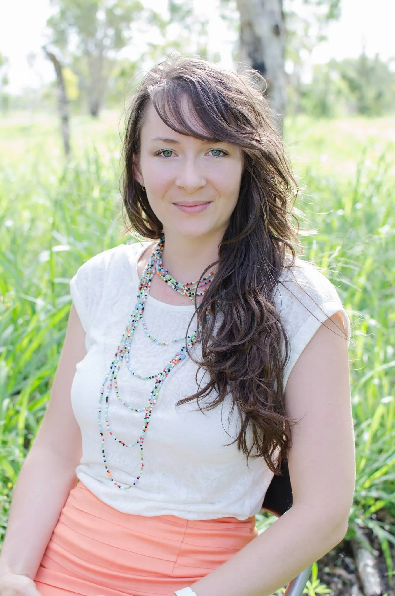 A woman with long, wavy brown hair and light green eyes, wearing a white lace top and coral-colored skirt, standing outdoors in a green, sunny setting with trees in the background.