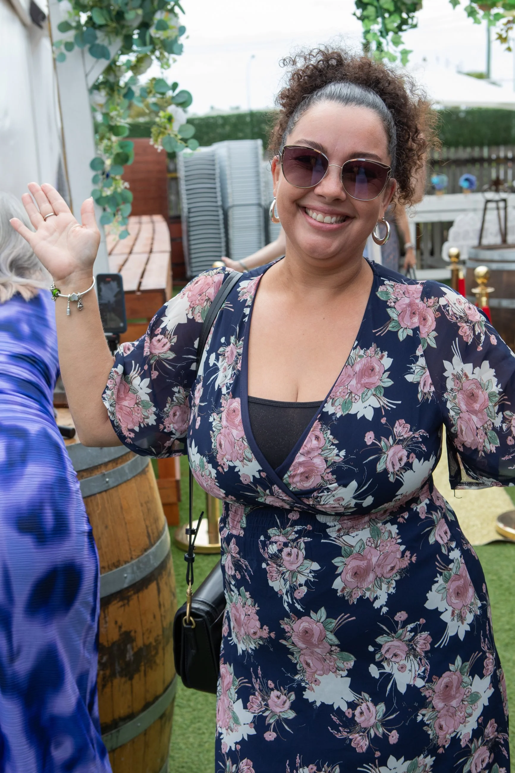 A smiling woman with curly hair, wearing sunglasses, earrings, and a floral dress, waving at a social gathering outdoors.