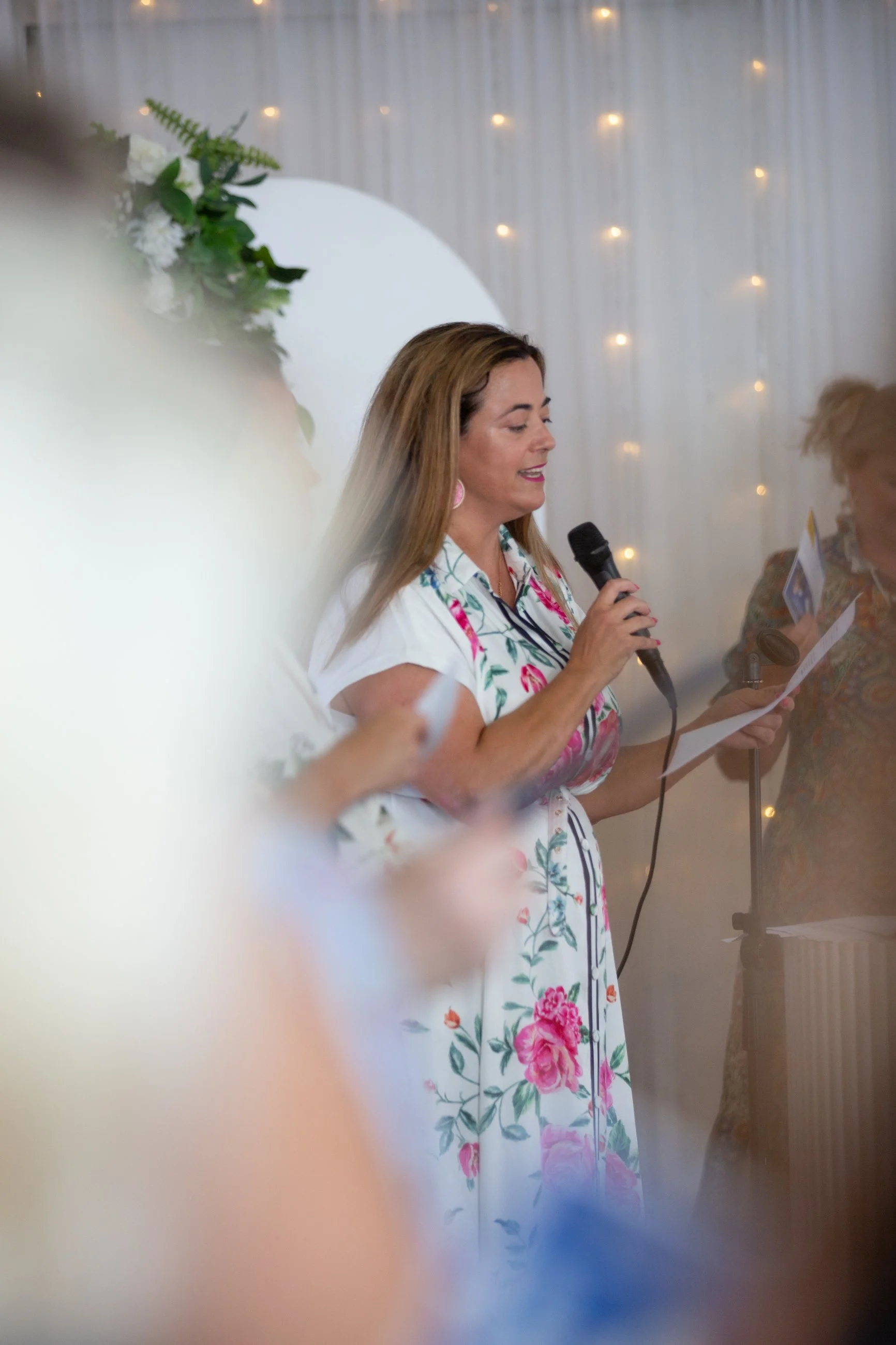 A woman with light brown hair wearing a white dress with pink and green floral patterns, holding a microphone and reading from a paper at a braless brunch event in brisbane with white fairy lights in the background.