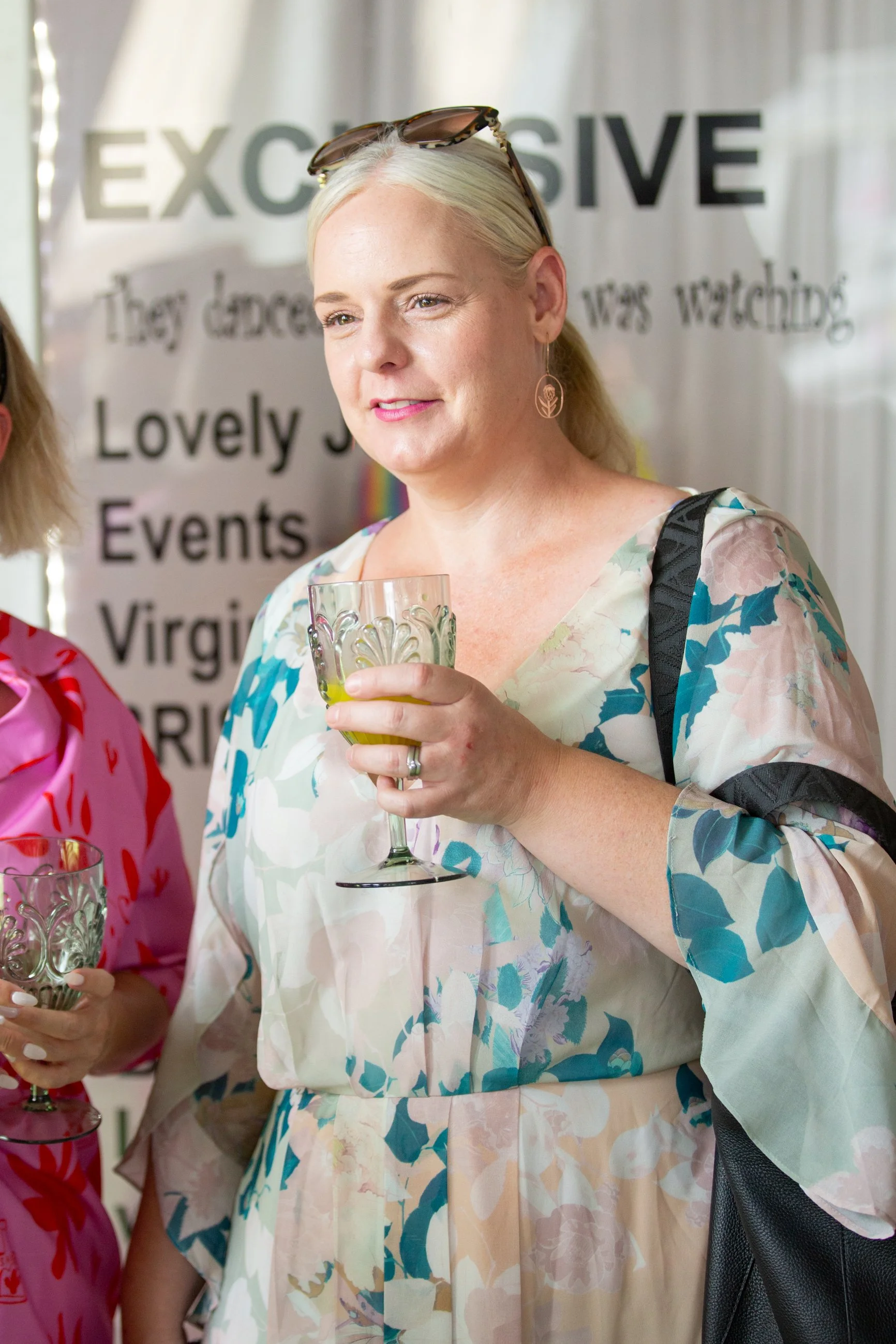 A woman with long blonde hair, wearing floral dress and sunglasses on her head, holding a glass of drink, smiling at a social braless brunch event.