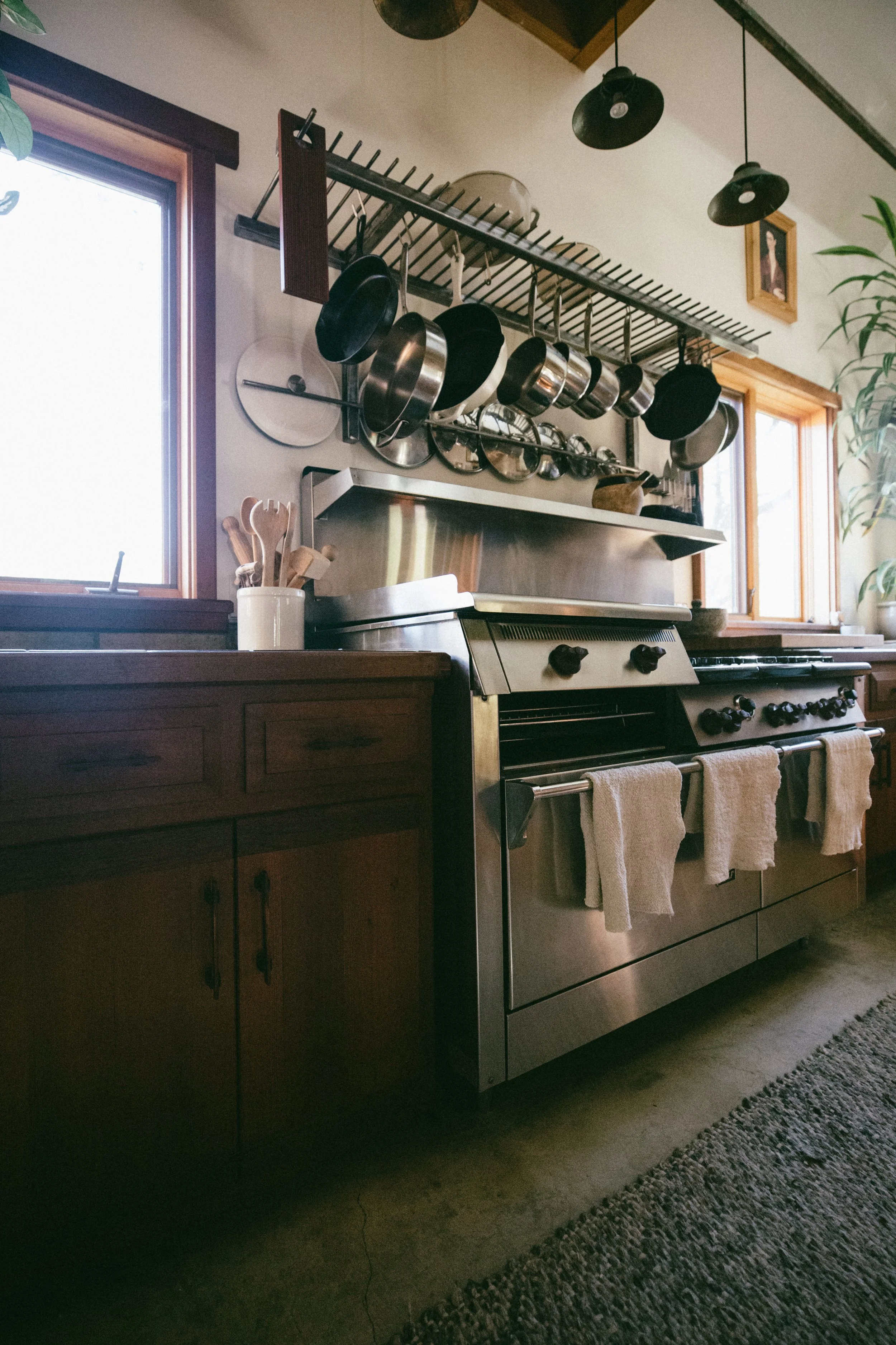 Modern kitchen with stainless steel stove, wooden counter, and hanging pots and pans above. Windows on either side let in natural light, and there are kitchen utensils in a white jar on the counter.