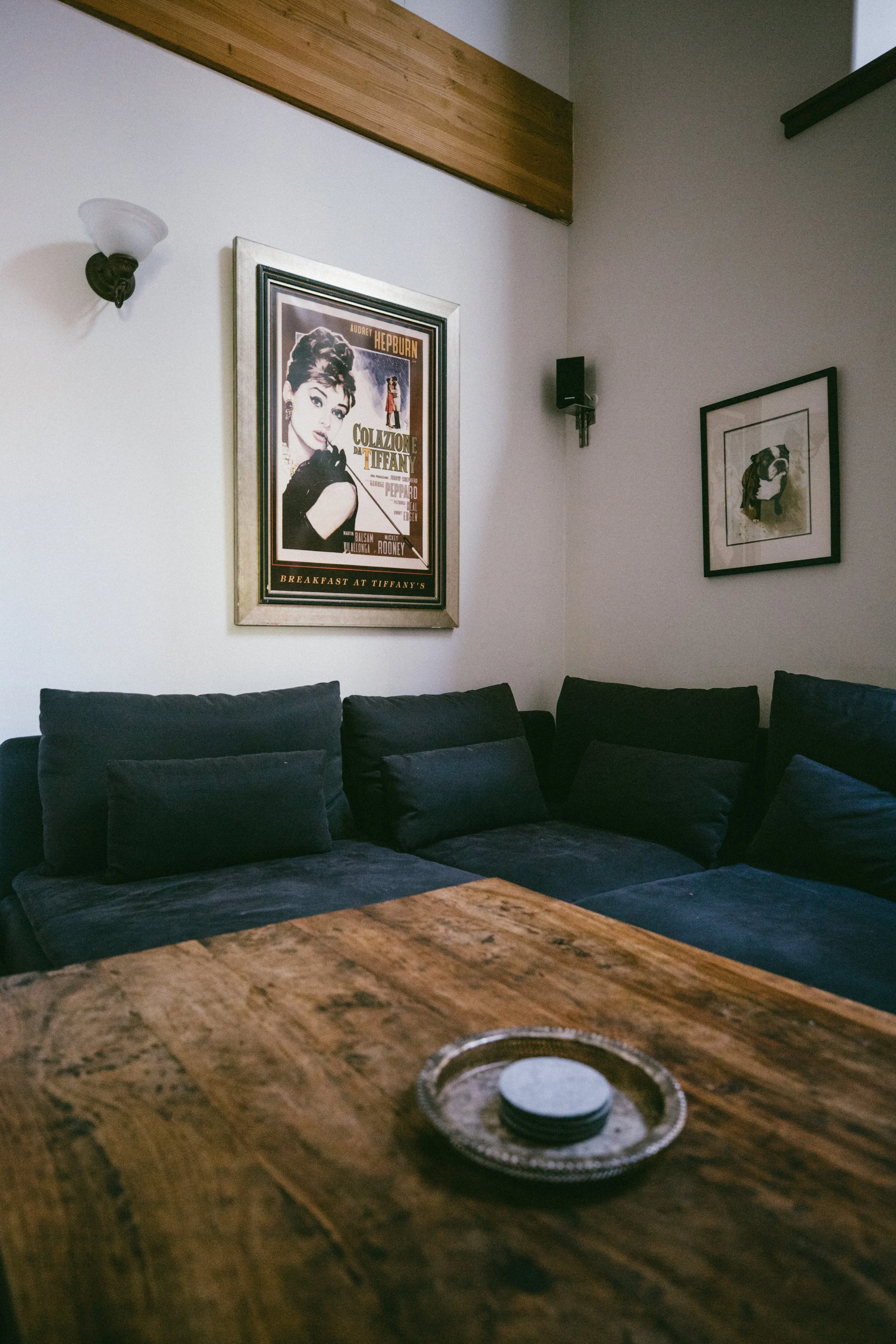 A cozy living room corner with a dark upholstered sectional sofa, a wooden coffee table with a round ashtray on it, framed vintage posters and artwork on the white walls, a wall-mounted speaker, and a wall-mounted light fixture.