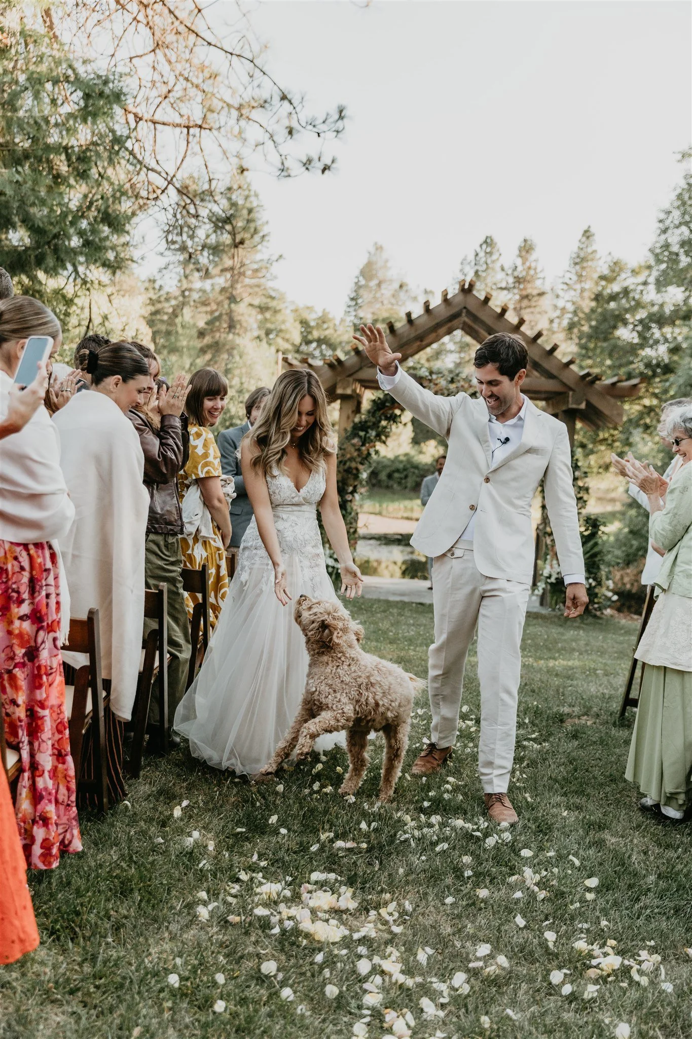 Bride and groom with a dog walking down an outdoor aisle at a wedding ceremony, guests clapping and taking photos, during daytime in a scenic outdoor setting.