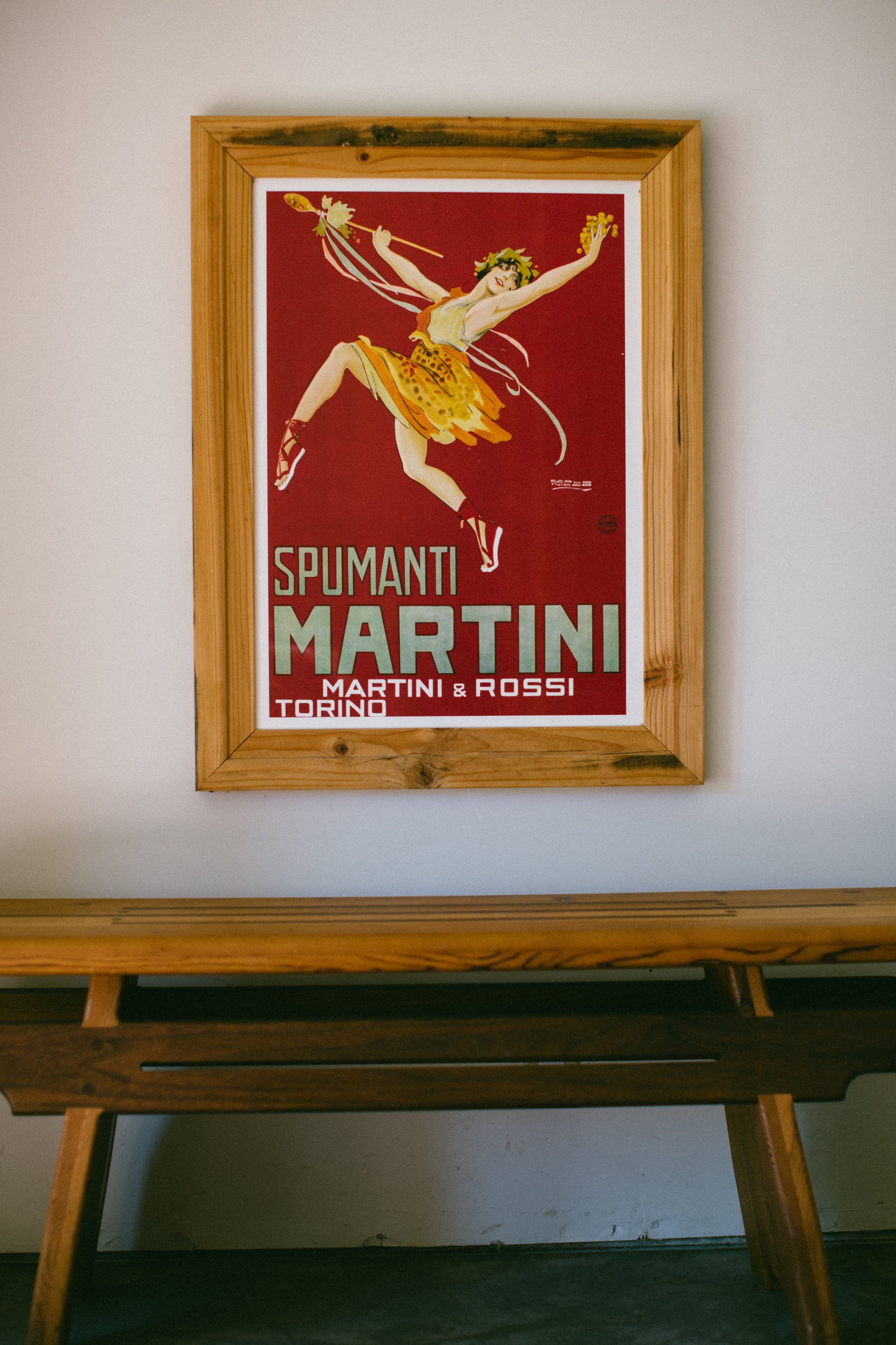 A framed vintage poster of a dancer in a yellow and orange costume with red shoes, jumping with one arm raised, on a red background. The poster advertises Spumanti Martini, Martini & Rossi, Torino.