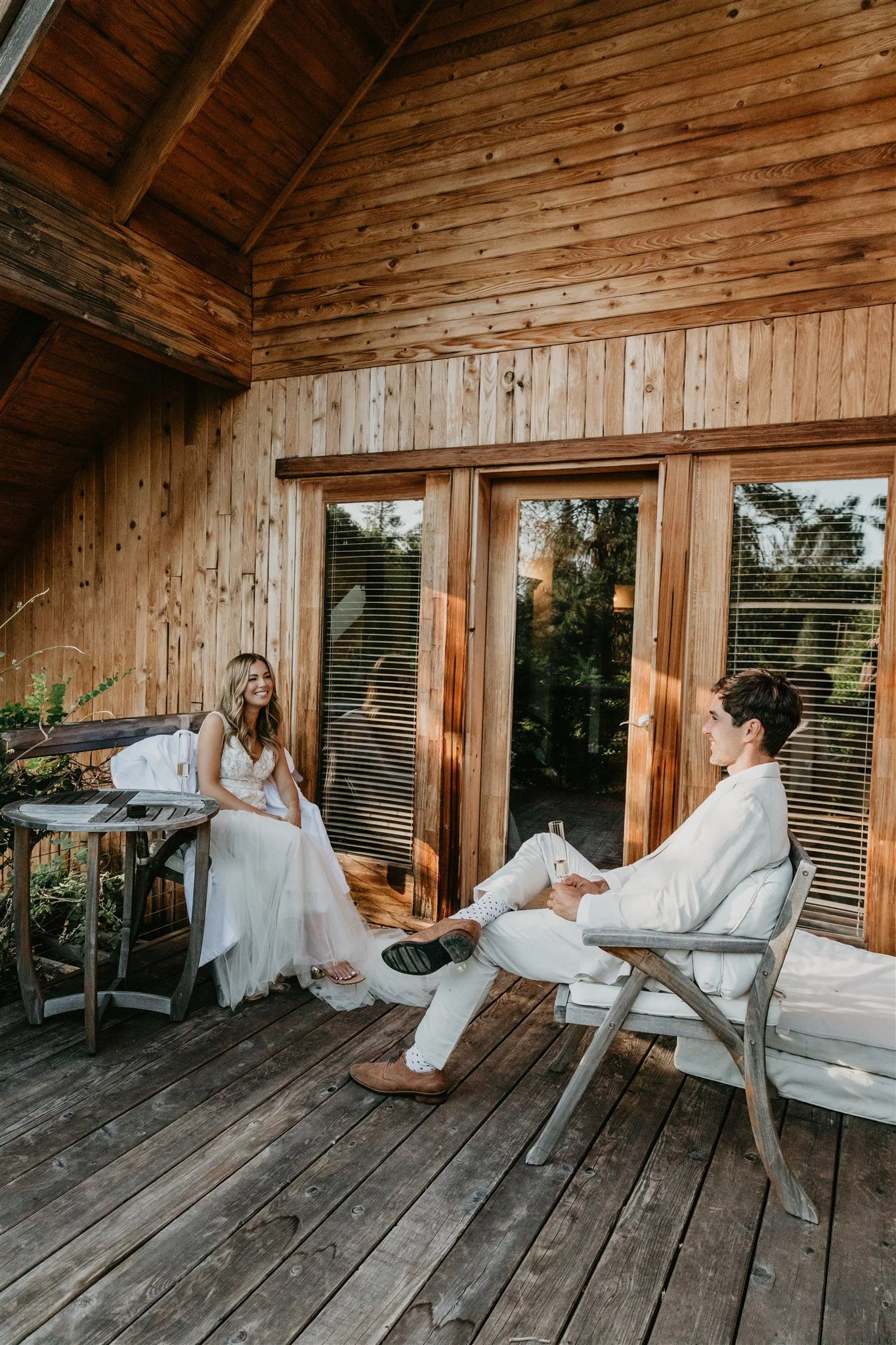 A woman in a wedding dress sitting on a chair, smiling, as a man in white attire sitting on a bench, holding a glass, talking to her on a wooden patio with large glass windows behind them.