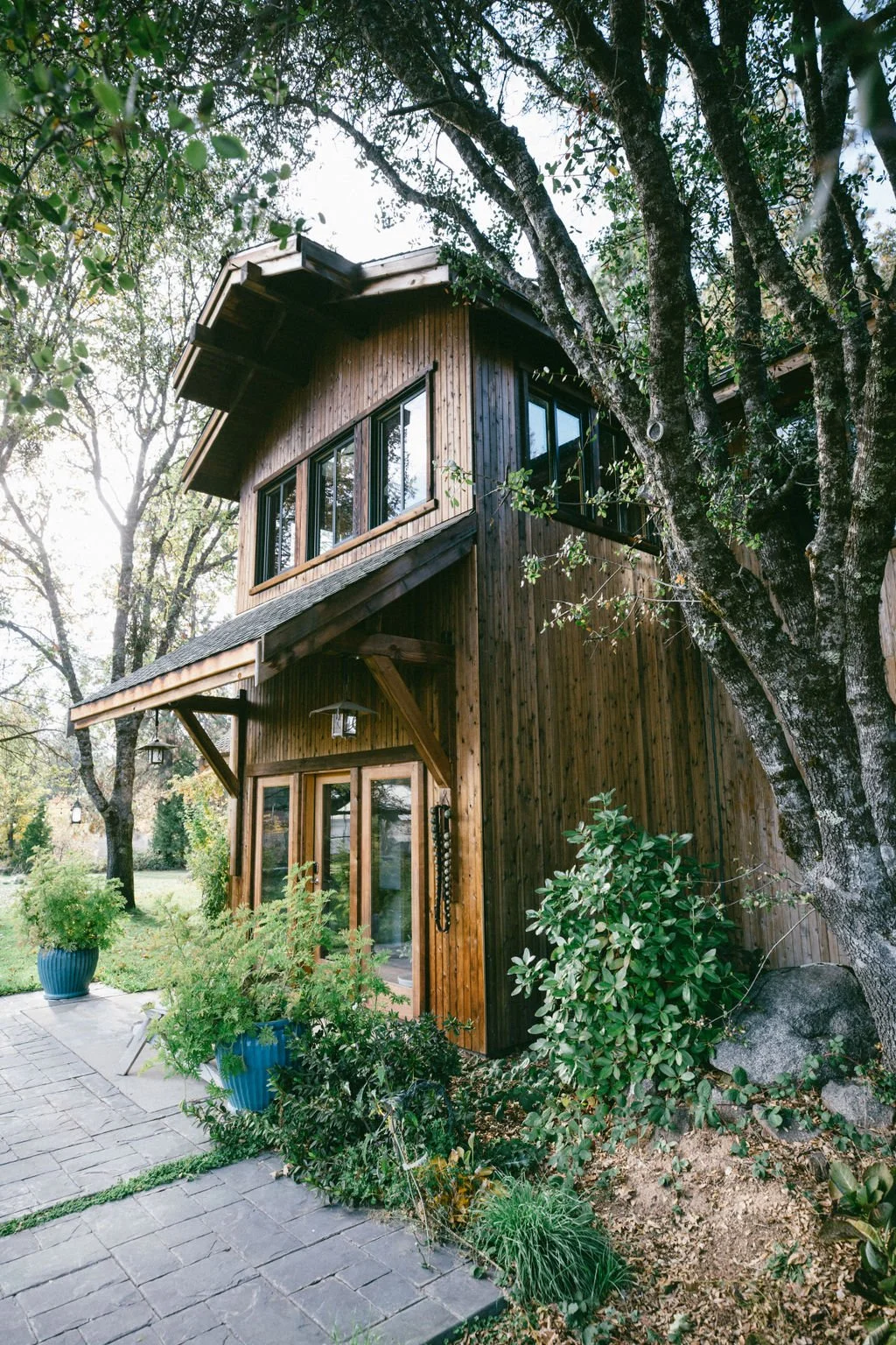 A two-story wooden house among trees with large windows, a stone pathway, and potted plants in blue containers.