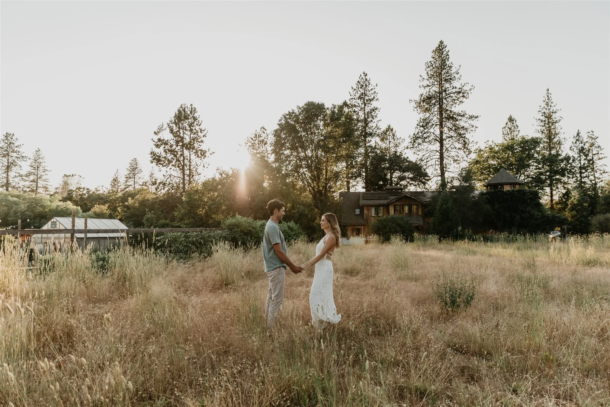 A couple holding hands in a grassy field during sunset, with trees and houses in the background.