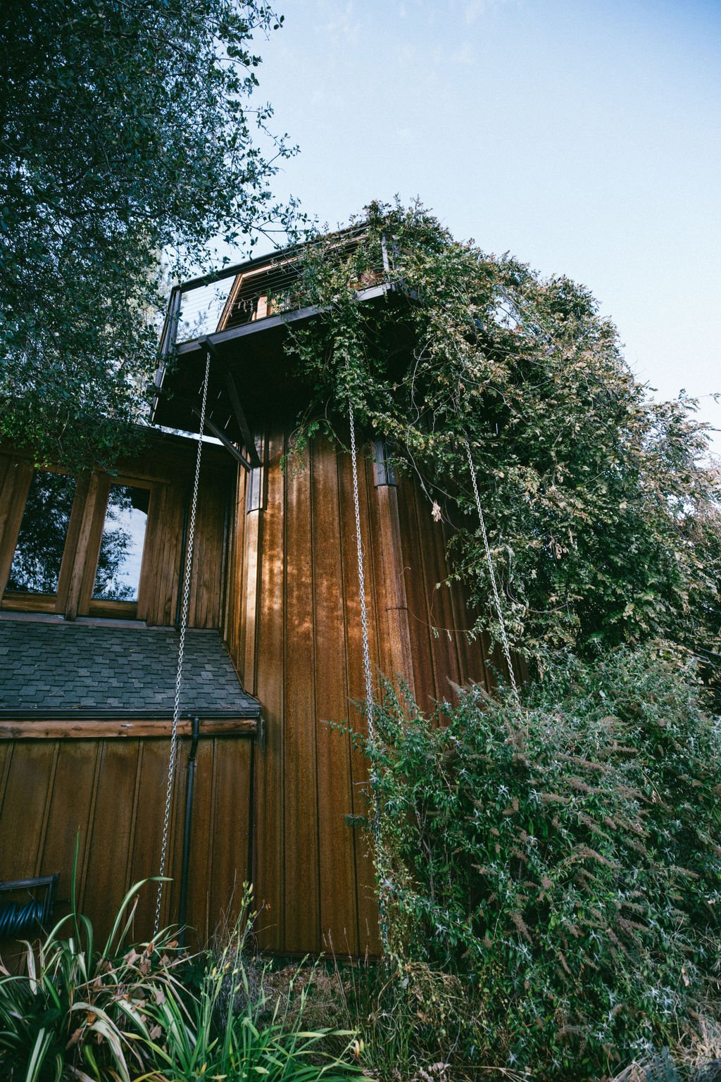 A tall wooden house with a tower, surrounded by trees and plants, with chains hanging from the upper balcony.