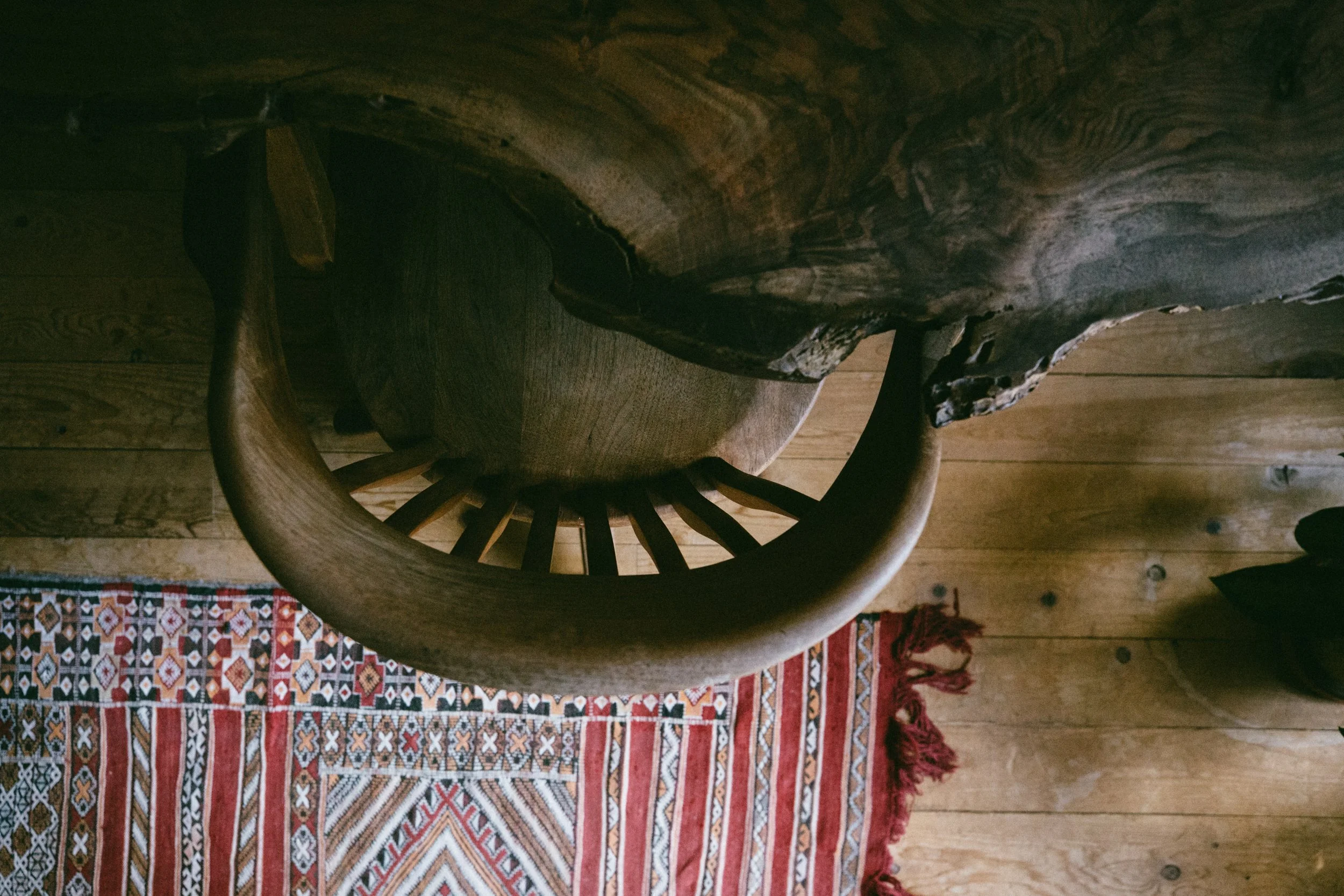 Top-down view of a wooden table with a round wooden seat underneath, next to a colorful woven rug on a wooden floor.