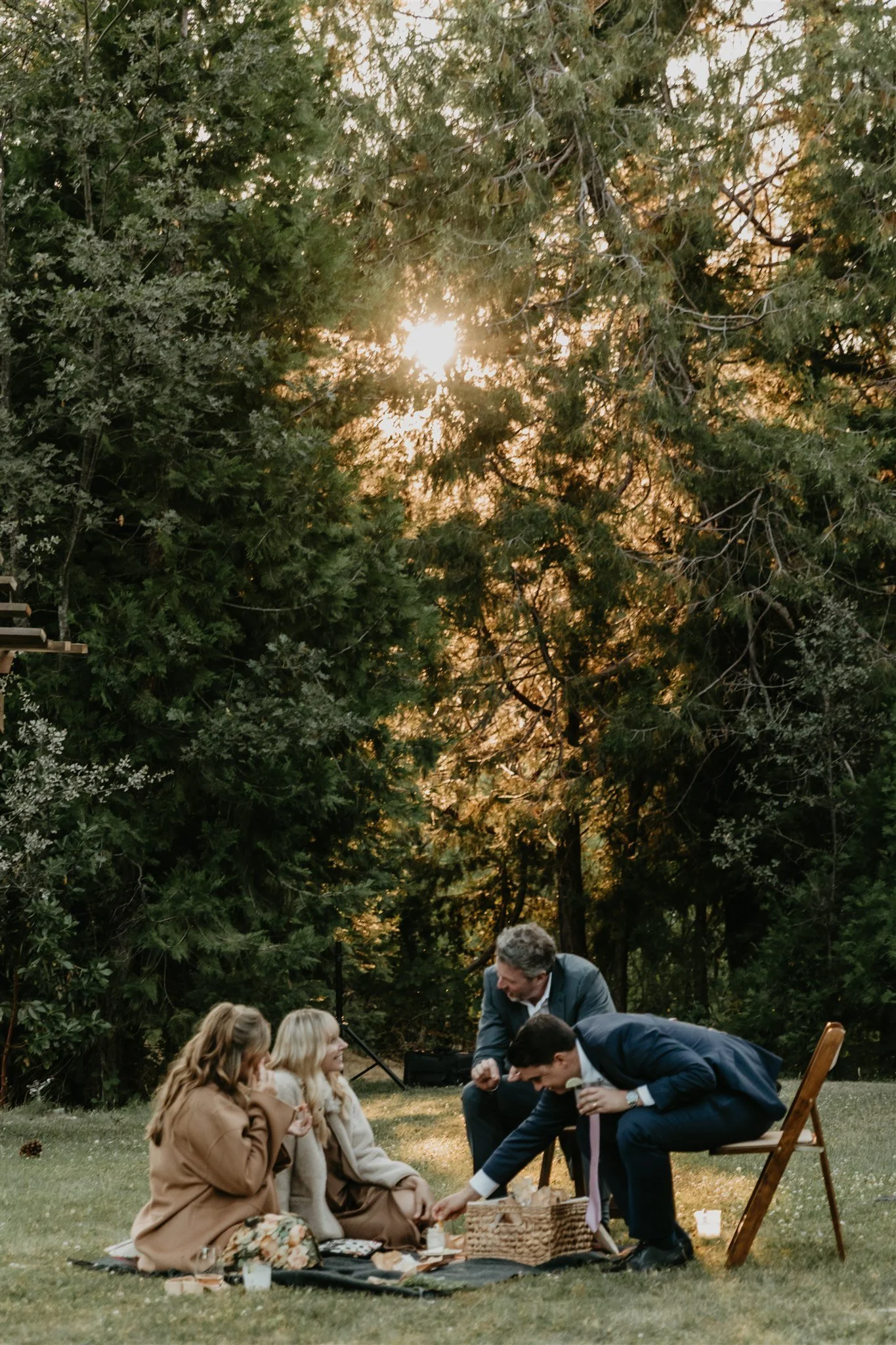 A group of five people having a picnic outdoors in a grassy area surrounded by tall trees during sunset. Two women and three men are smiling, chatting, and sharing food on a blanket with a picnic basket.