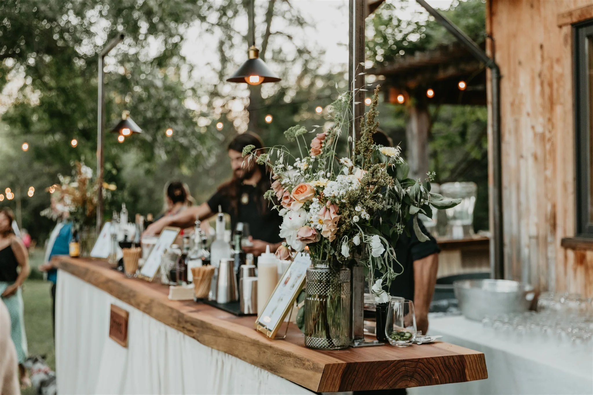 Outdoor wedding bar with a large bouquet of flowers in a glass vase, surrounded by bottles and drink accessories, during dusk with string lights and trees in the background.