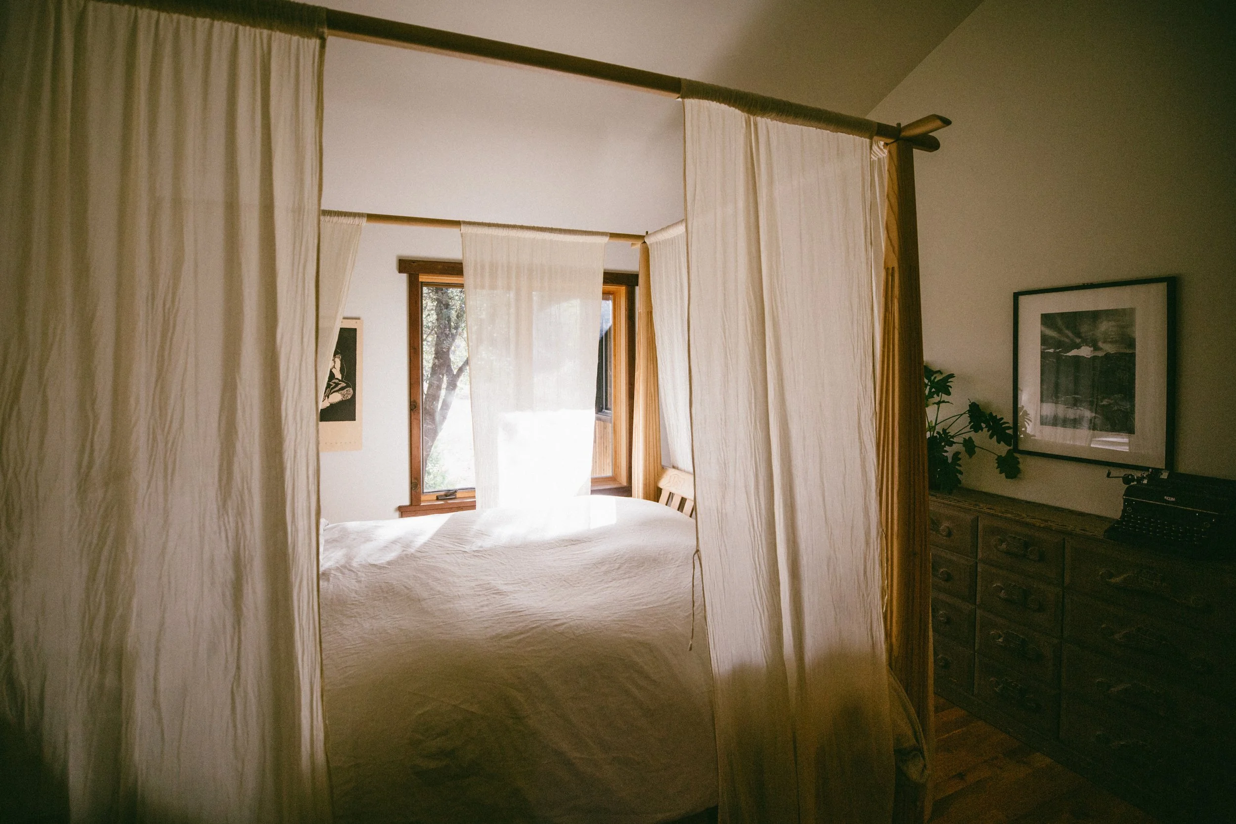 A cozy bedroom with a bed under a canopy of cream-colored curtains, sunlight streaming through a wooden-framed window, and a wooden dresser with framed artwork hanging above it.