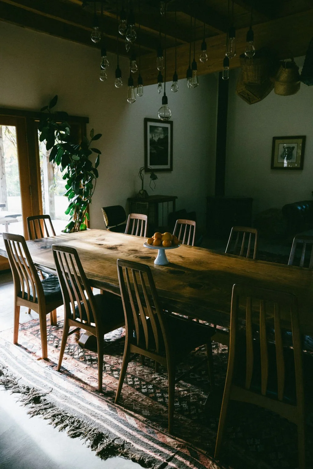 A dining room with a large wooden table surrounded by wooden chairs, a fruit centerpiece, a patterned rug, and hanging light bulbs. There is a sliding glass door with a plant beside it and framed pictures on the walls.