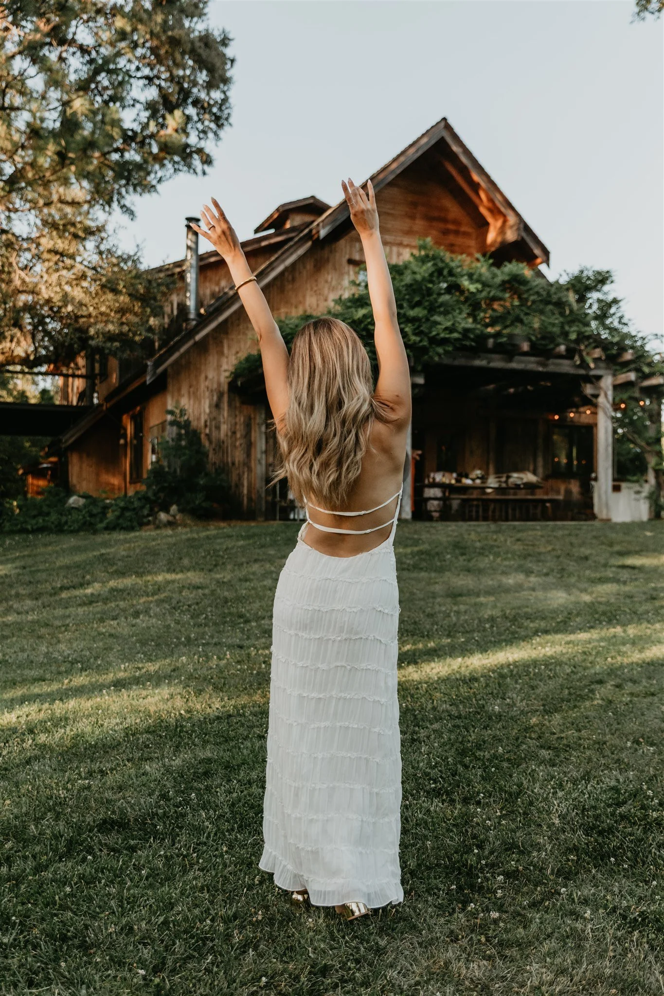 A woman with long blonde hair wearing a white strapless dress standing on a grassy lawn with her arms raised in front of a rustic wooden house.