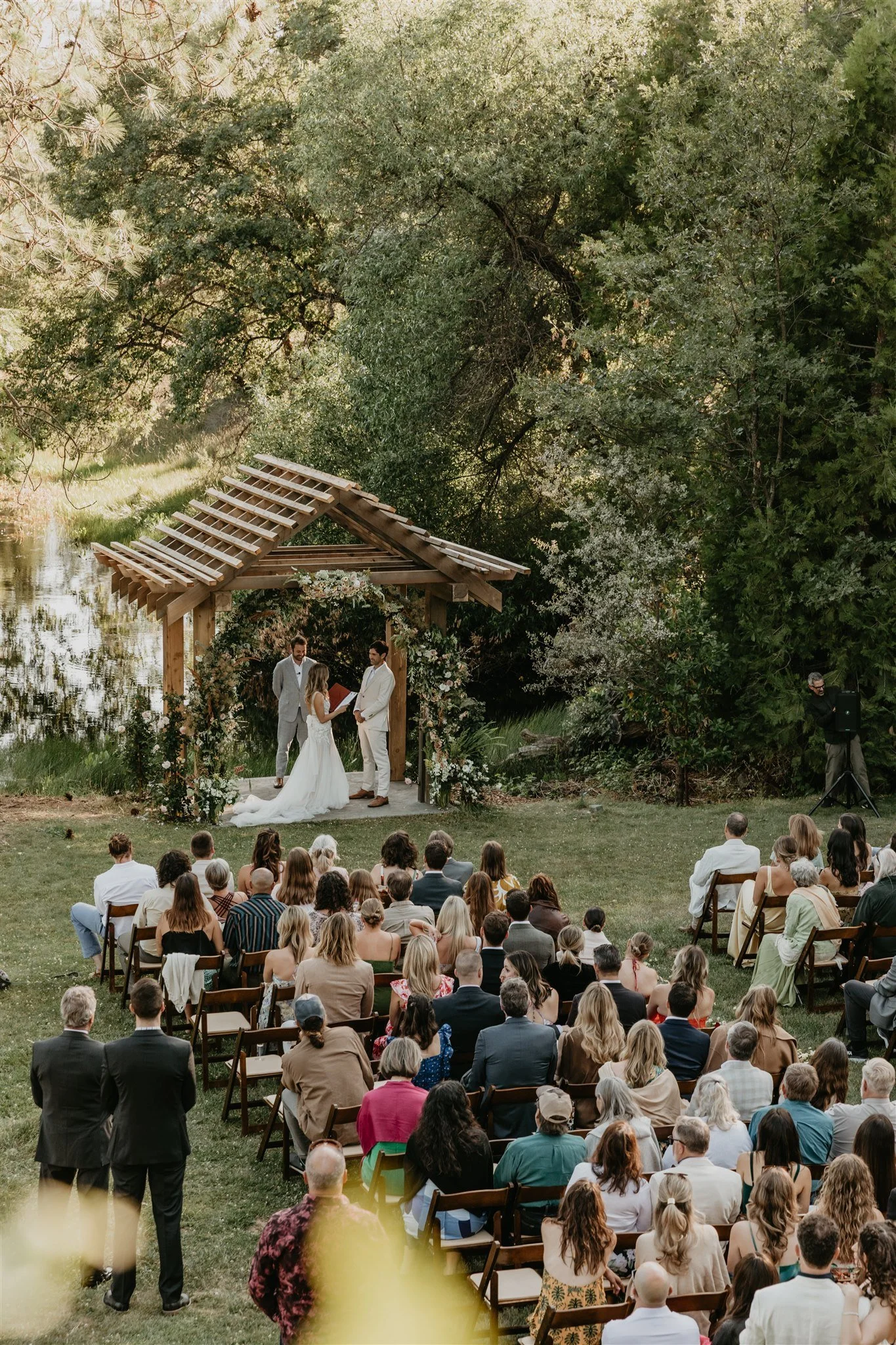 A wedding ceremony taking place outdoors under a wooden arch decorated with flowers, surrounded by trees and guests seated on chairs, with a couple exchanging vows and an officiant in front.