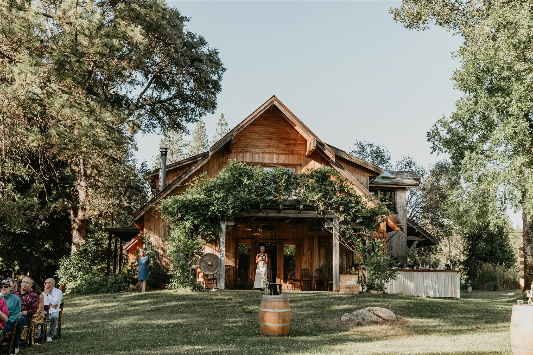 A rustic wooden house with front porch surrounded by large trees and greenery. There is an outdoor gathering with chairs on the lawn, and a woman in white near the entrance.